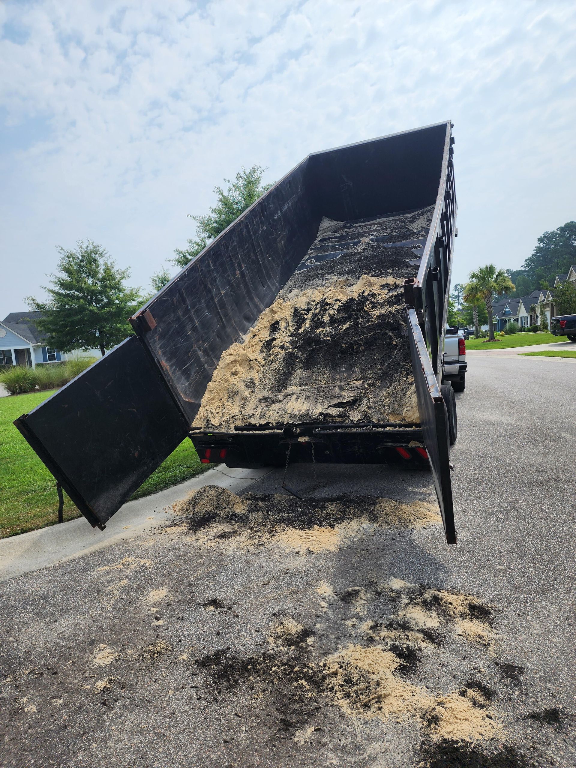 A black dump truck with its bed raised, spewing wood chips onto a paved road in a residential area.
