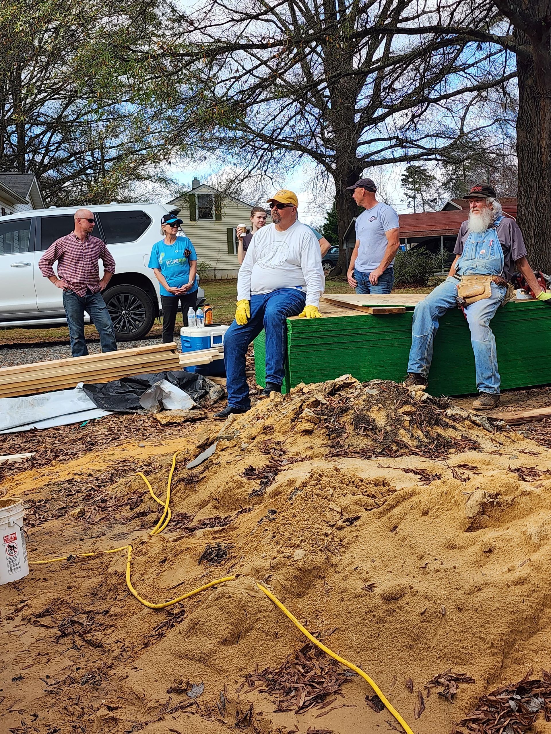 Group of people building outside, likely a house.  Men and women standing around wood materials, some wearing work gloves and tools.