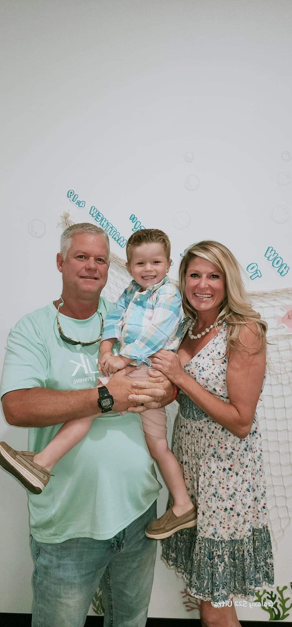 A family of three poses in front of a white wall. A man holds a young boy, while a woman stands beside them. They are smiling.