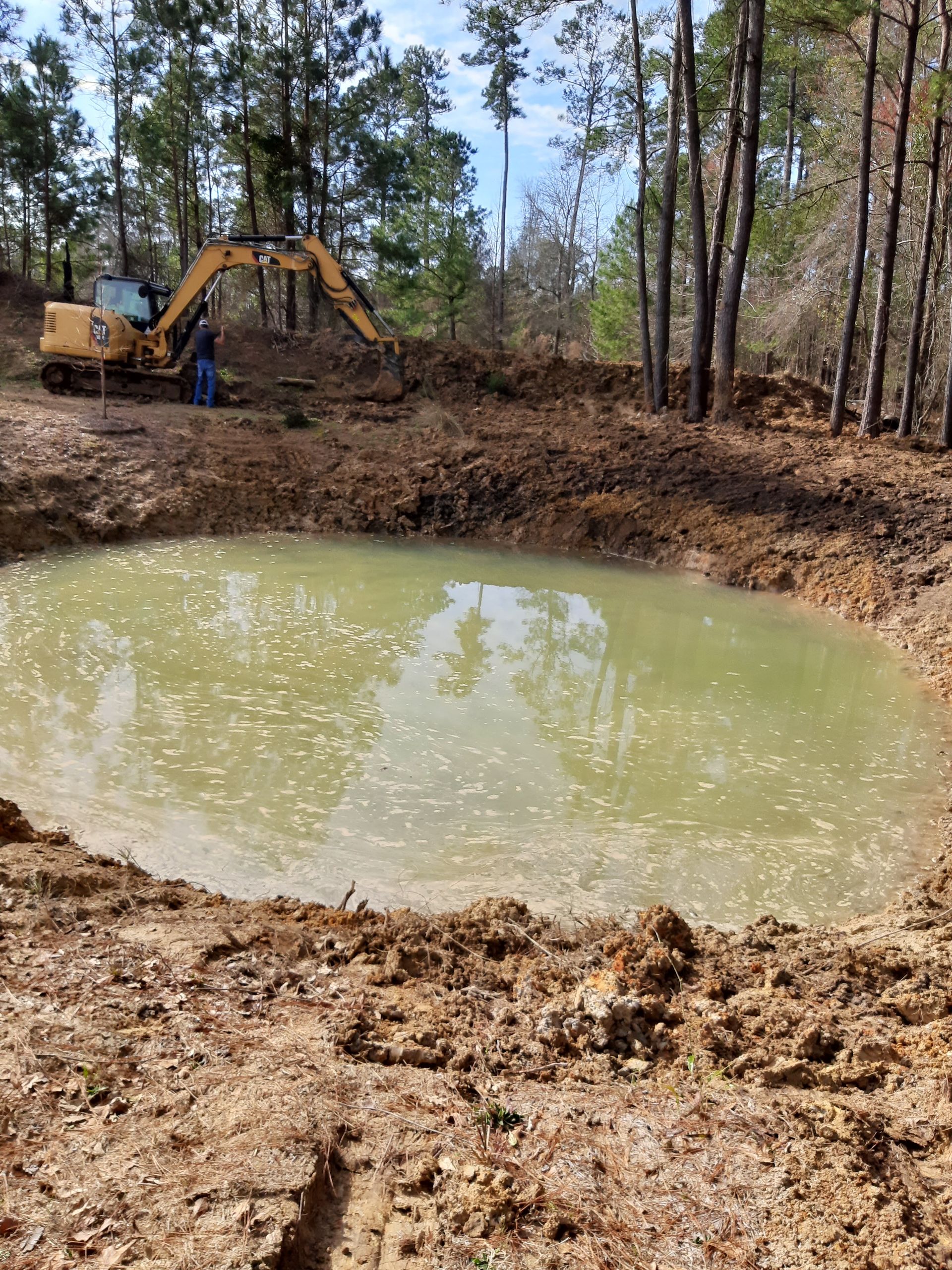 A newly dug pond filled with murky water, with a yellow excavator in the background and trees around the perimeter.