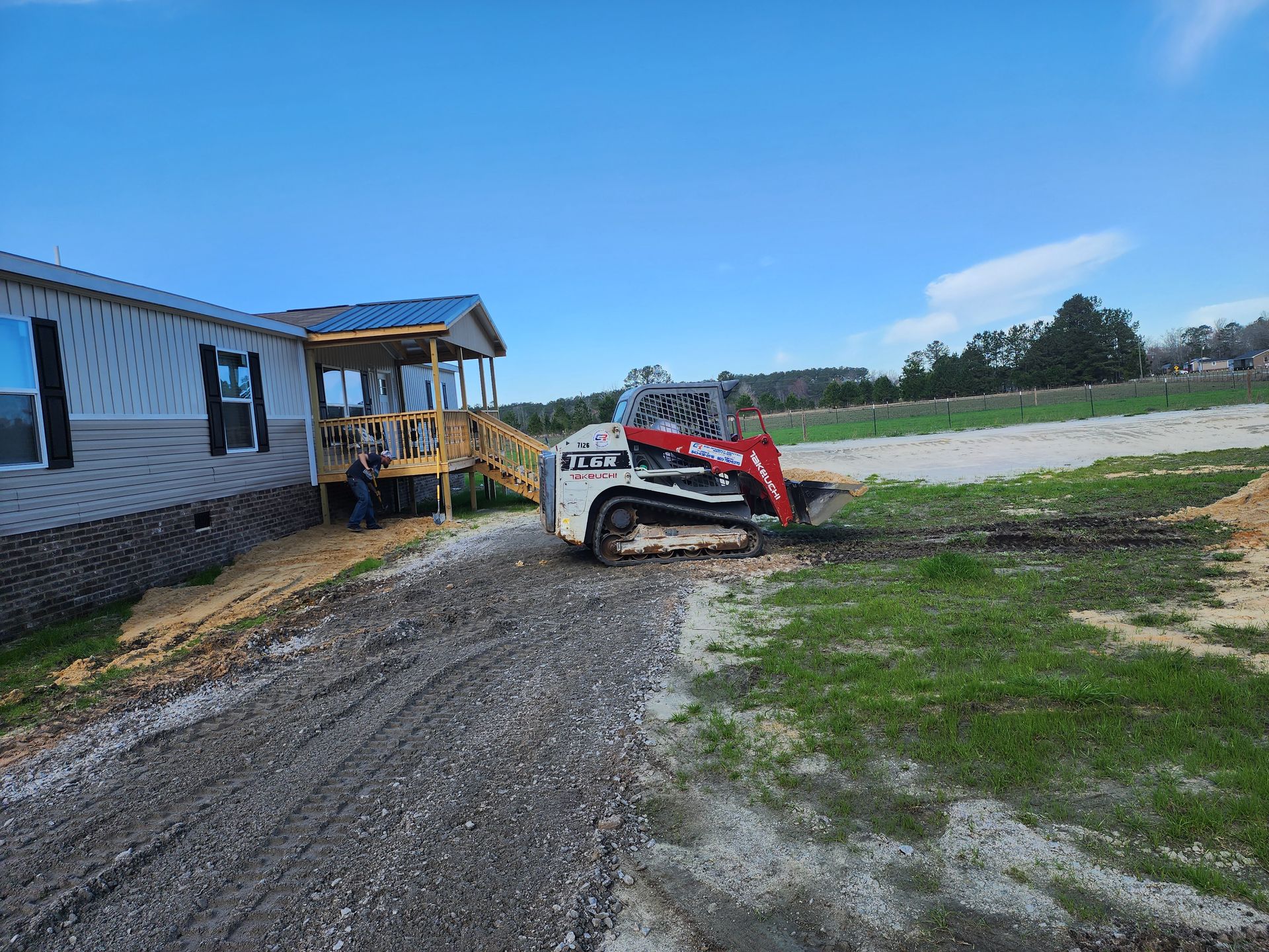 A white and red skid steer operating on a gravel driveway next to a manufactured home with a wooden porch under a blue sky.