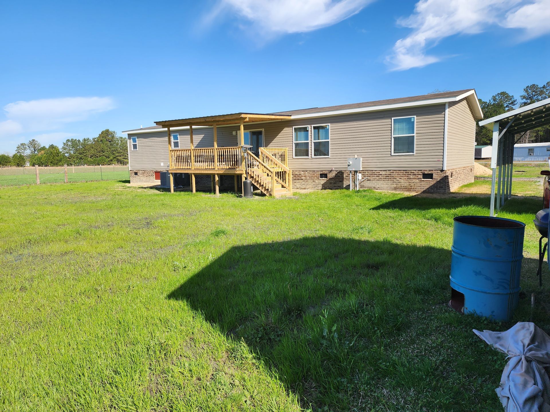 A single-story home with tan siding, a wooden deck, and a grassy yard under a bright blue sky.
