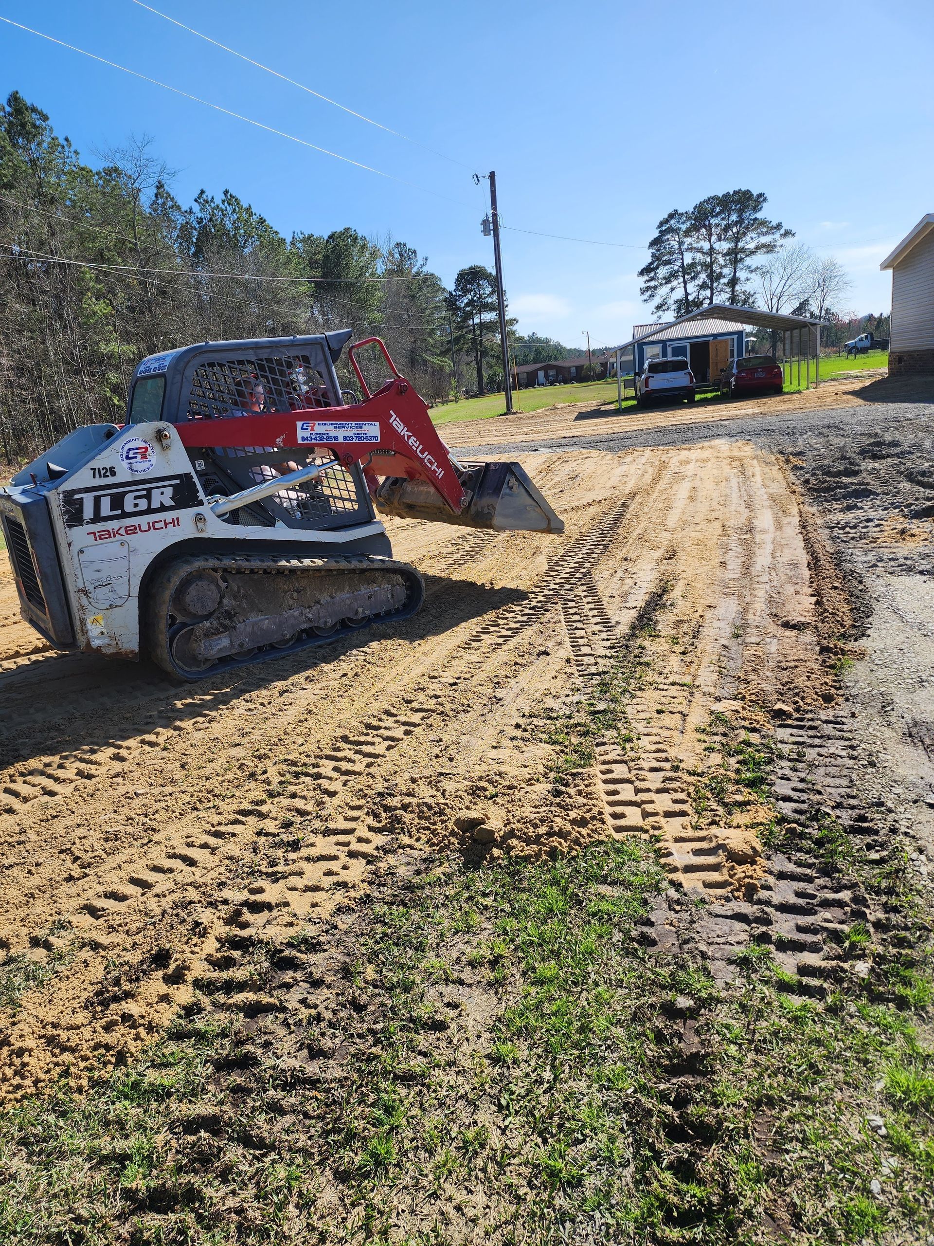 A white and red track loader is grading a sandy area next to a building in a sunny outdoor setting.