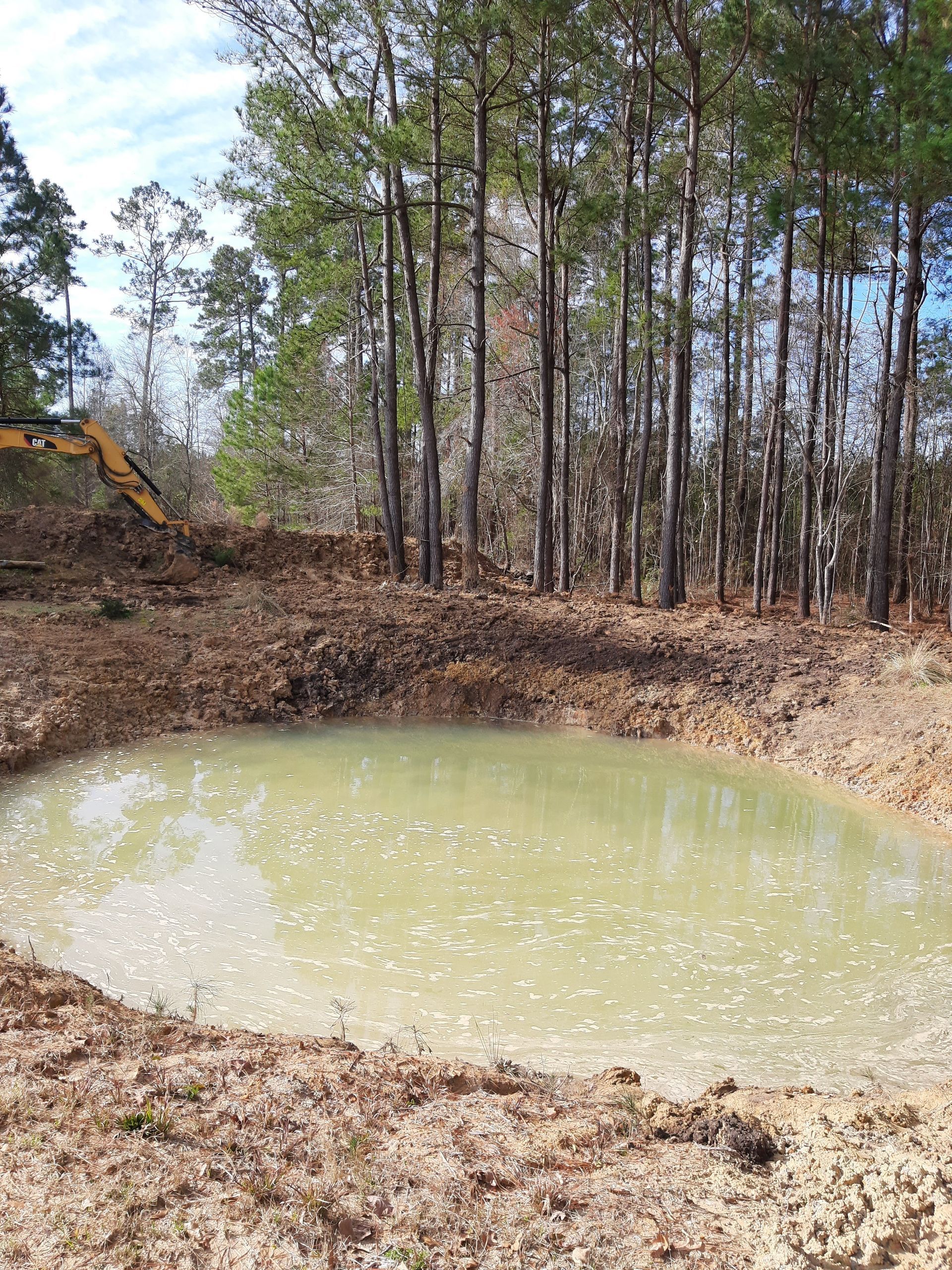 A newly dug pond filled with murky water sits in front of a line of tall trees; a yellow excavator is visible.