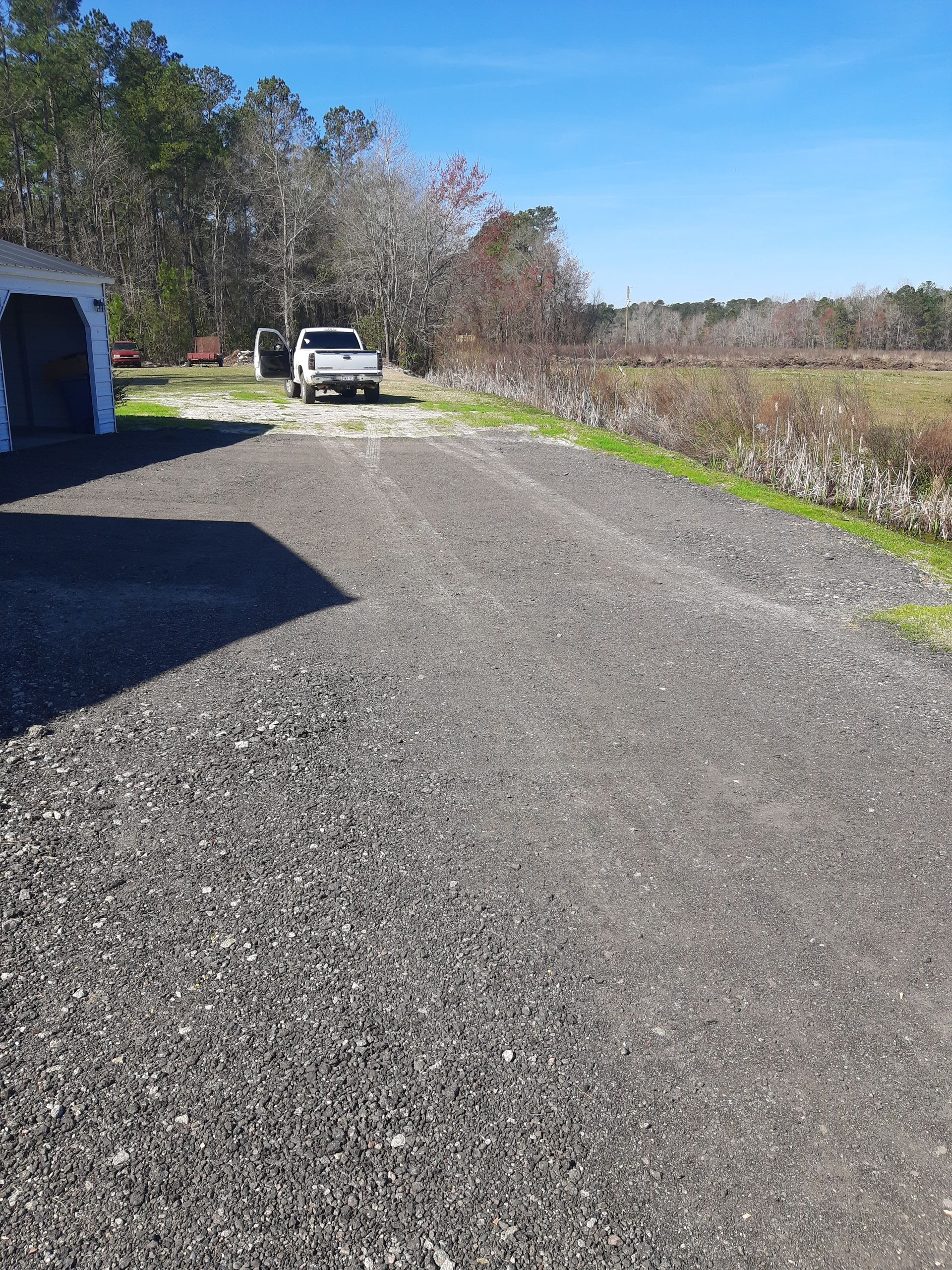 Gravel driveway with a white pickup truck parked in front of a field and some trees under a blue sky.