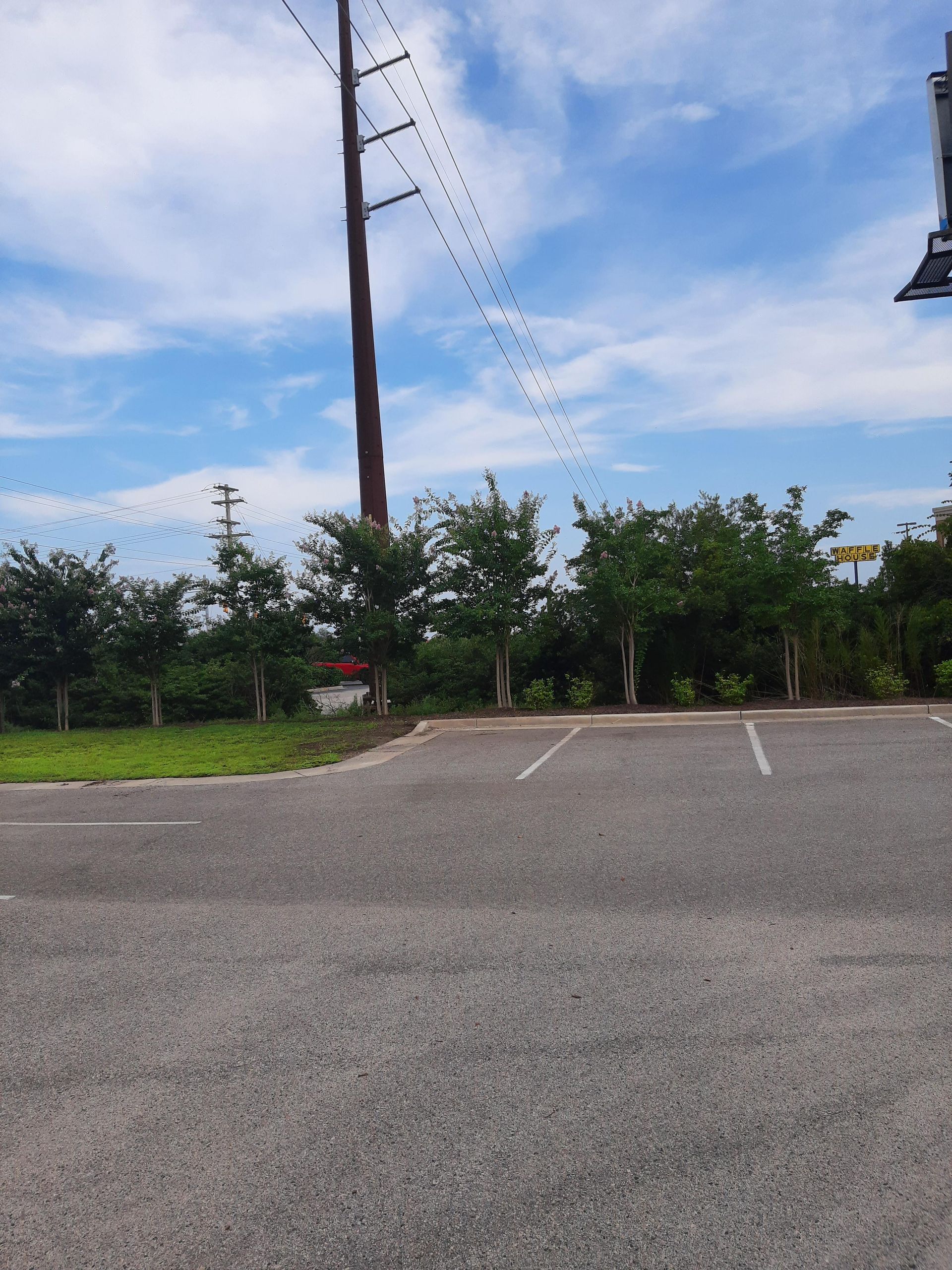 Empty parking lot with a row of trees, power lines, and a blue sky with clouds in the background.