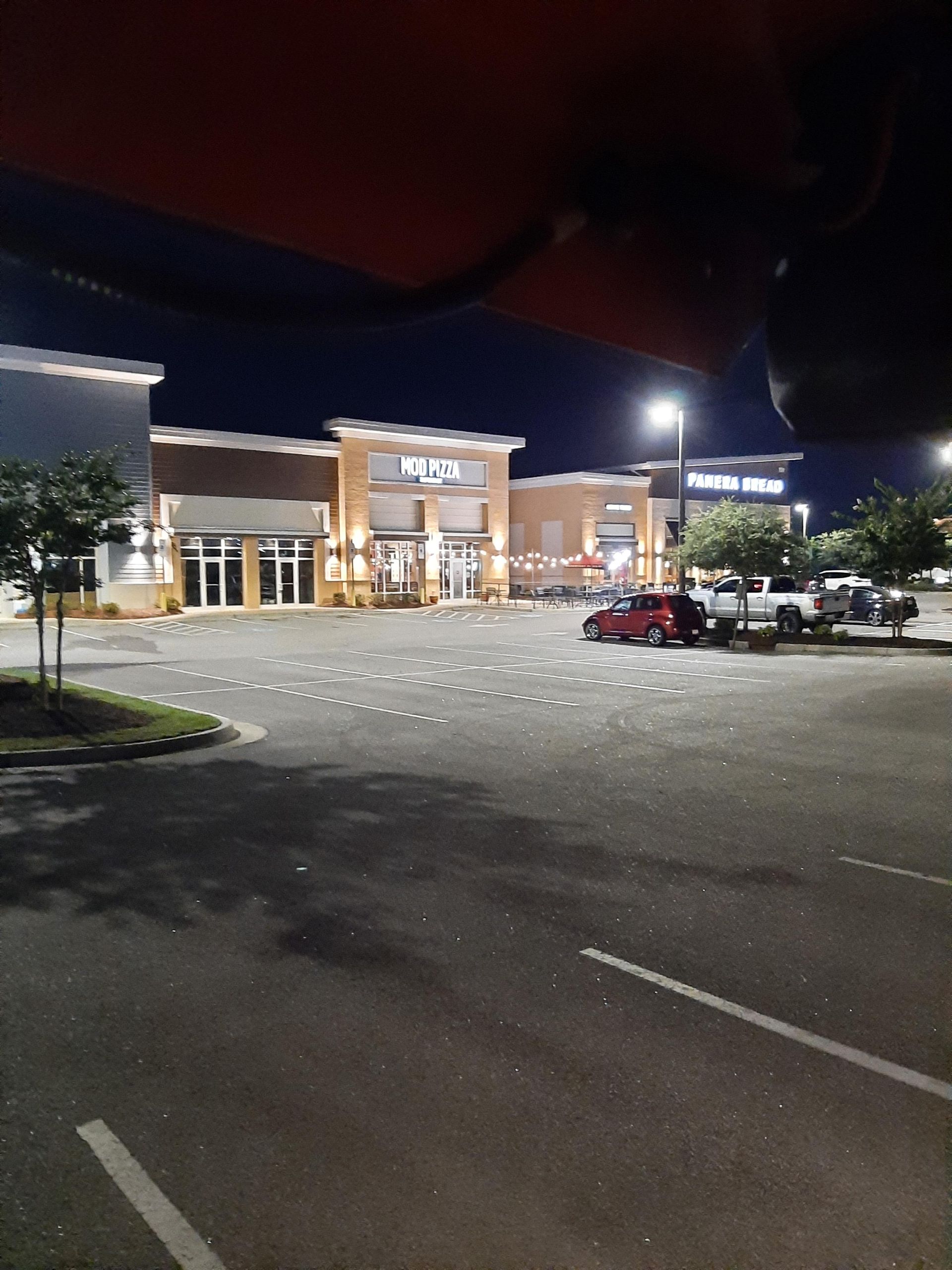Nighttime view of a shopping center with a partially lit parking lot and storefronts. A red car is parked near the buildings.