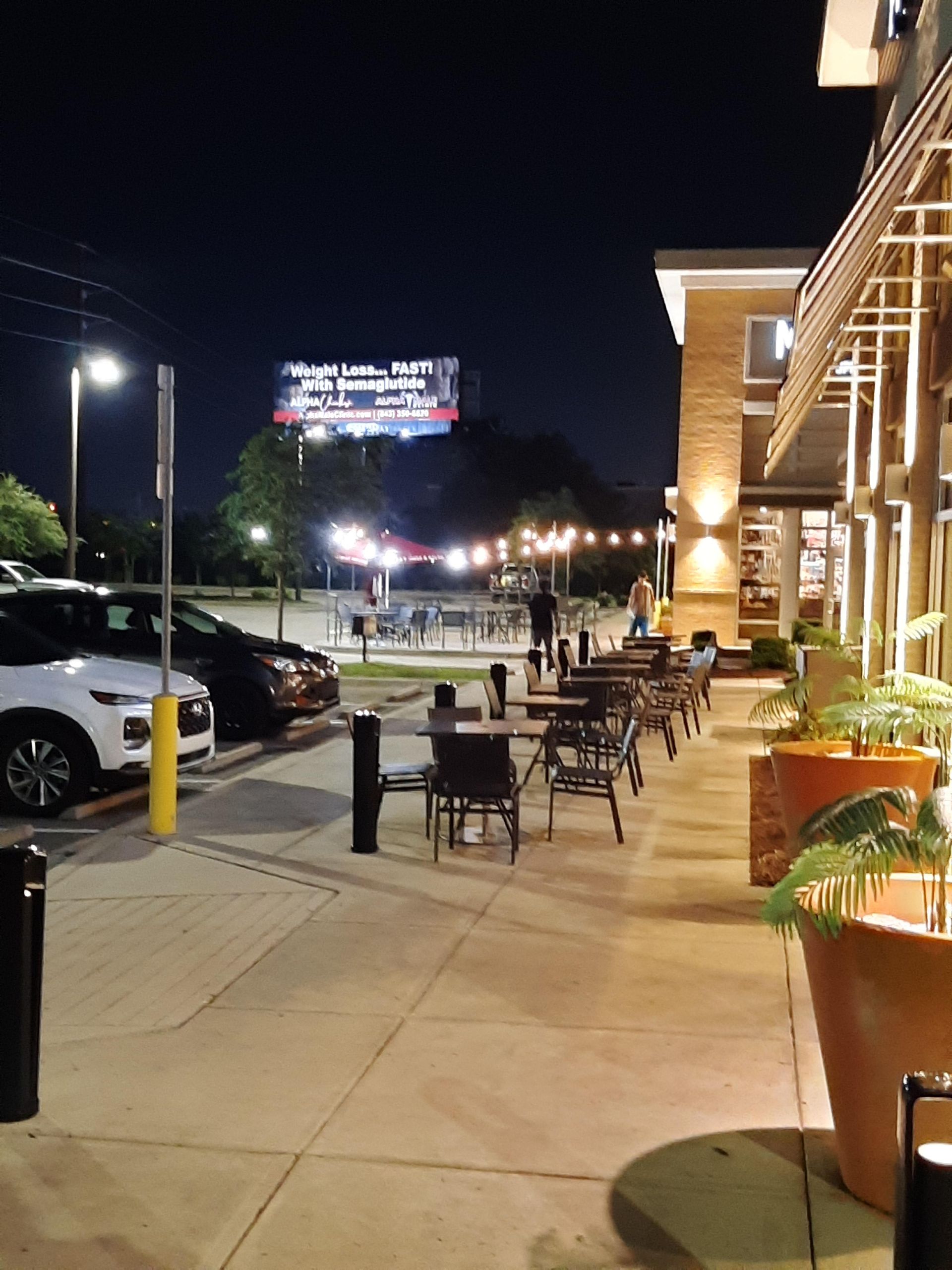 Nighttime outdoor dining area with tables, string lights, and parked cars. A billboard is visible in the background.