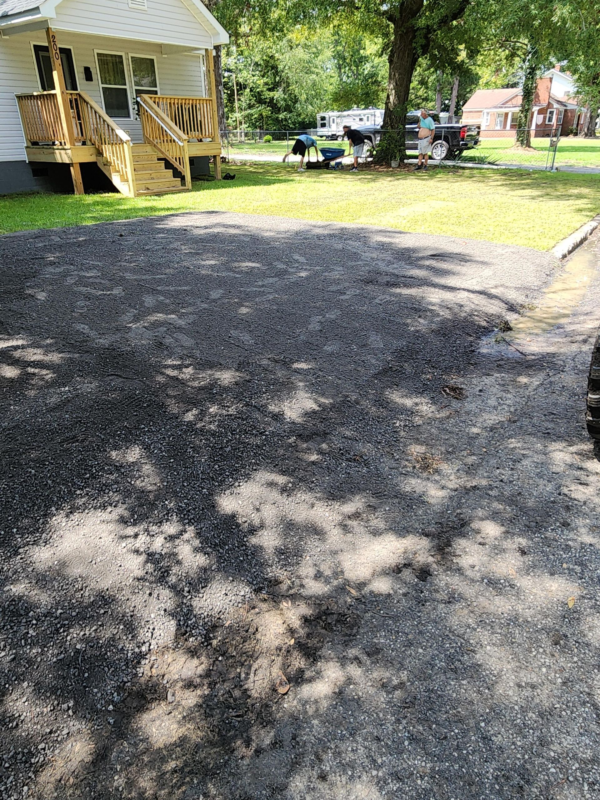Gravel driveway in front of a small house with a wooden porch and steps, tree shadow dappling the surface.