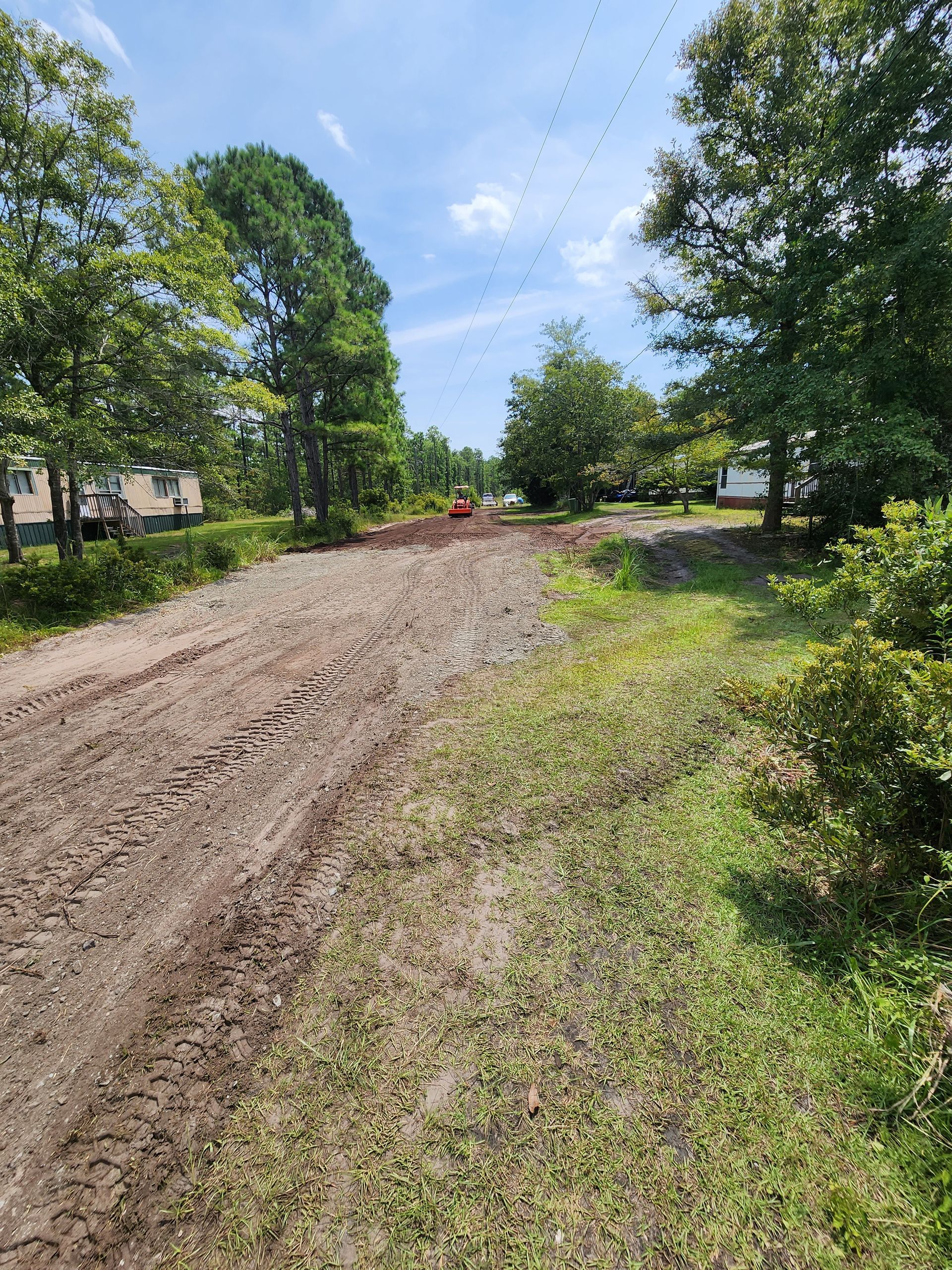Muddy dirt road with exposed rocks, trees on either side, under a blue sky. A small tractor is visible in the distance.