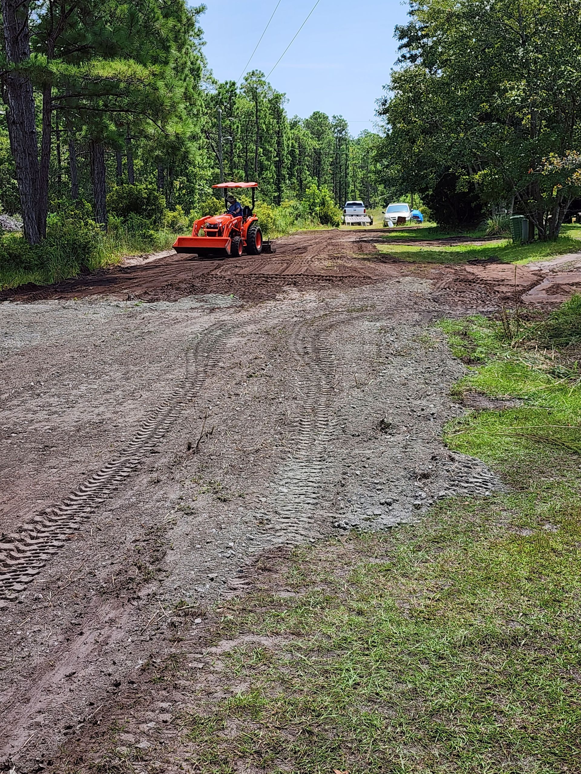 A tractor levels gravel on a muddy dirt road under a sunny sky, with trees on either side.