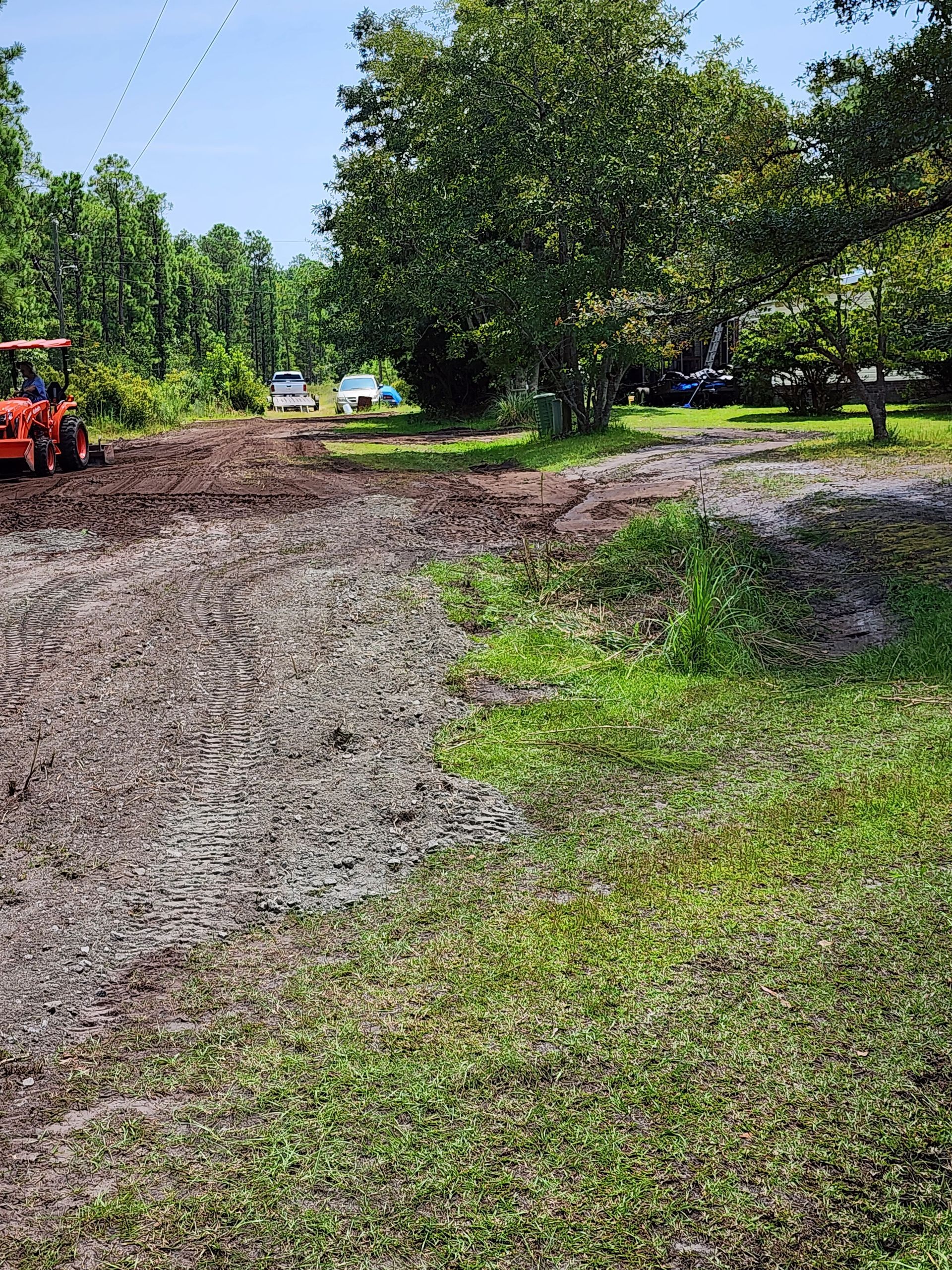 A dirt driveway with a tractor, muddy patches, and grassy areas under a sunny sky. Trees line the background.