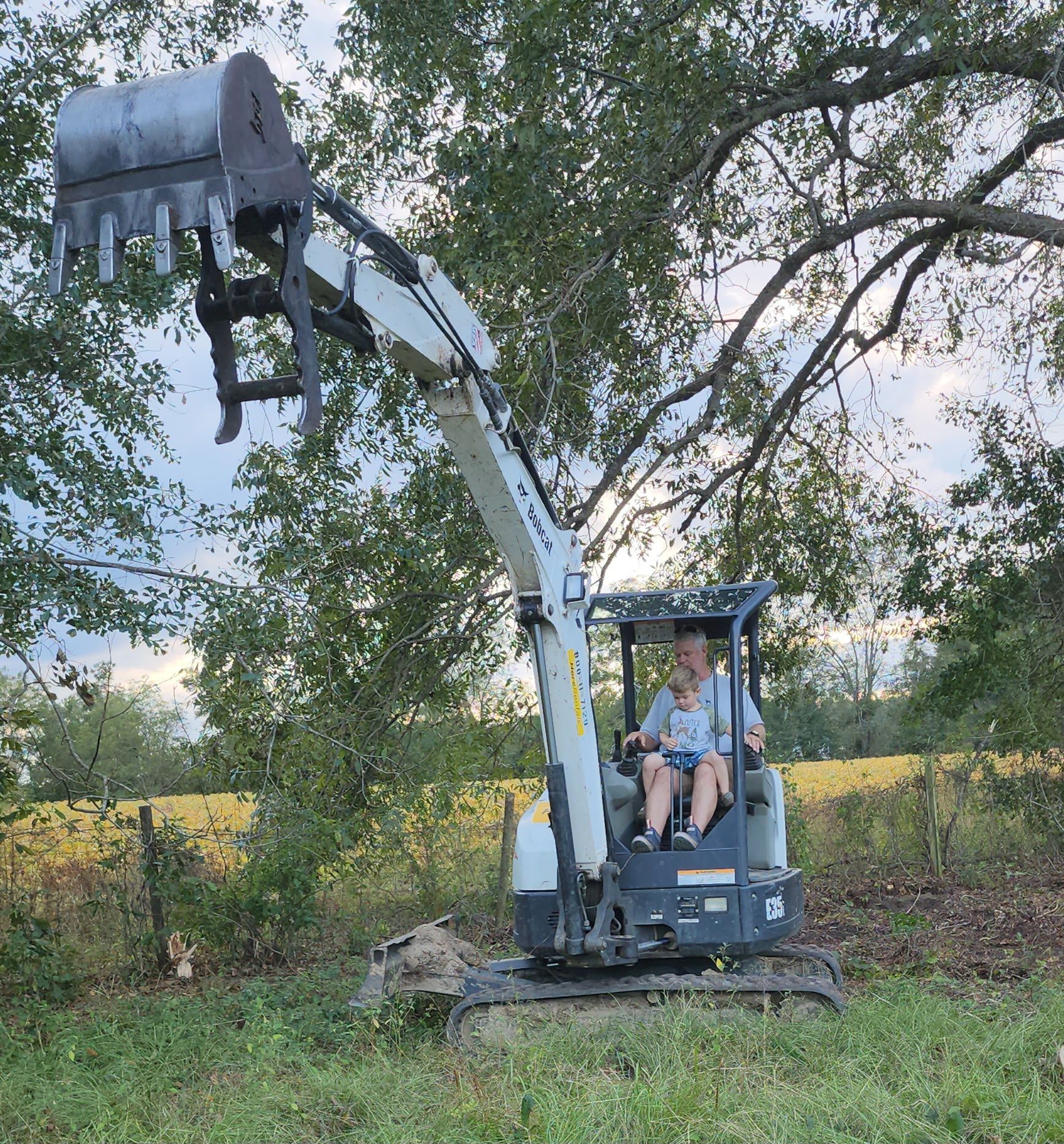 A person operates a small excavator with a grapple attachment outdoors. The machine is white and gray, and the setting is grassy with trees.
