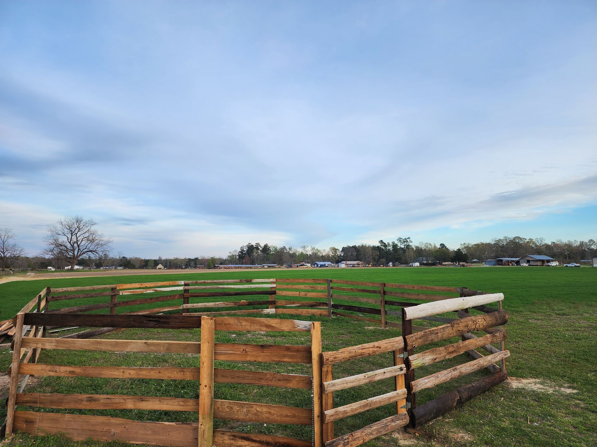 Wooden fence in a field under a cloudy blue sky. Distant trees and houses are visible on the horizon.