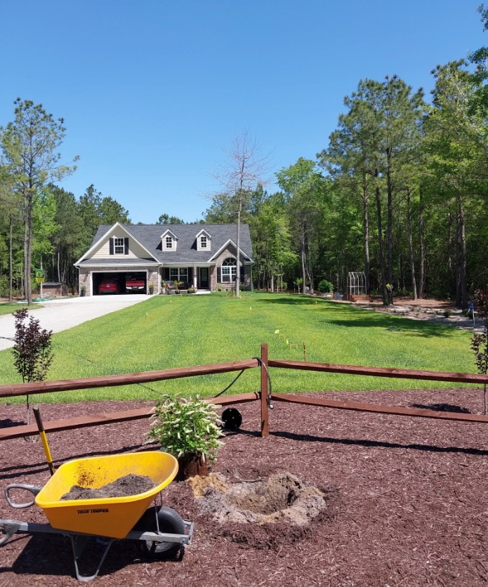 A house with a green lawn, trees, and a brown fence. A yellow wheelbarrow sits in the foreground with landscaping work underway.