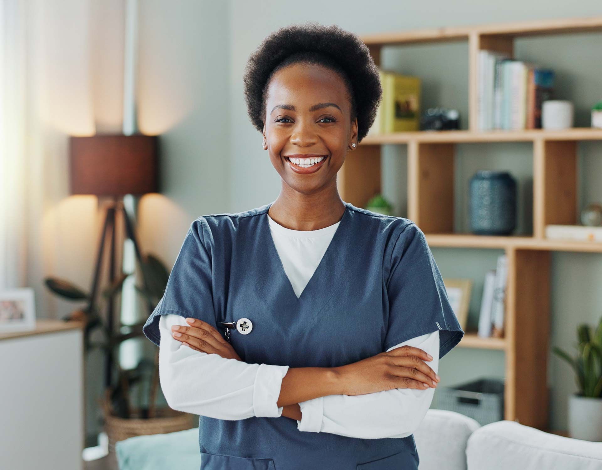 Smiling person in blue scrubs with arms crossed, standing in a room with a bookcase.