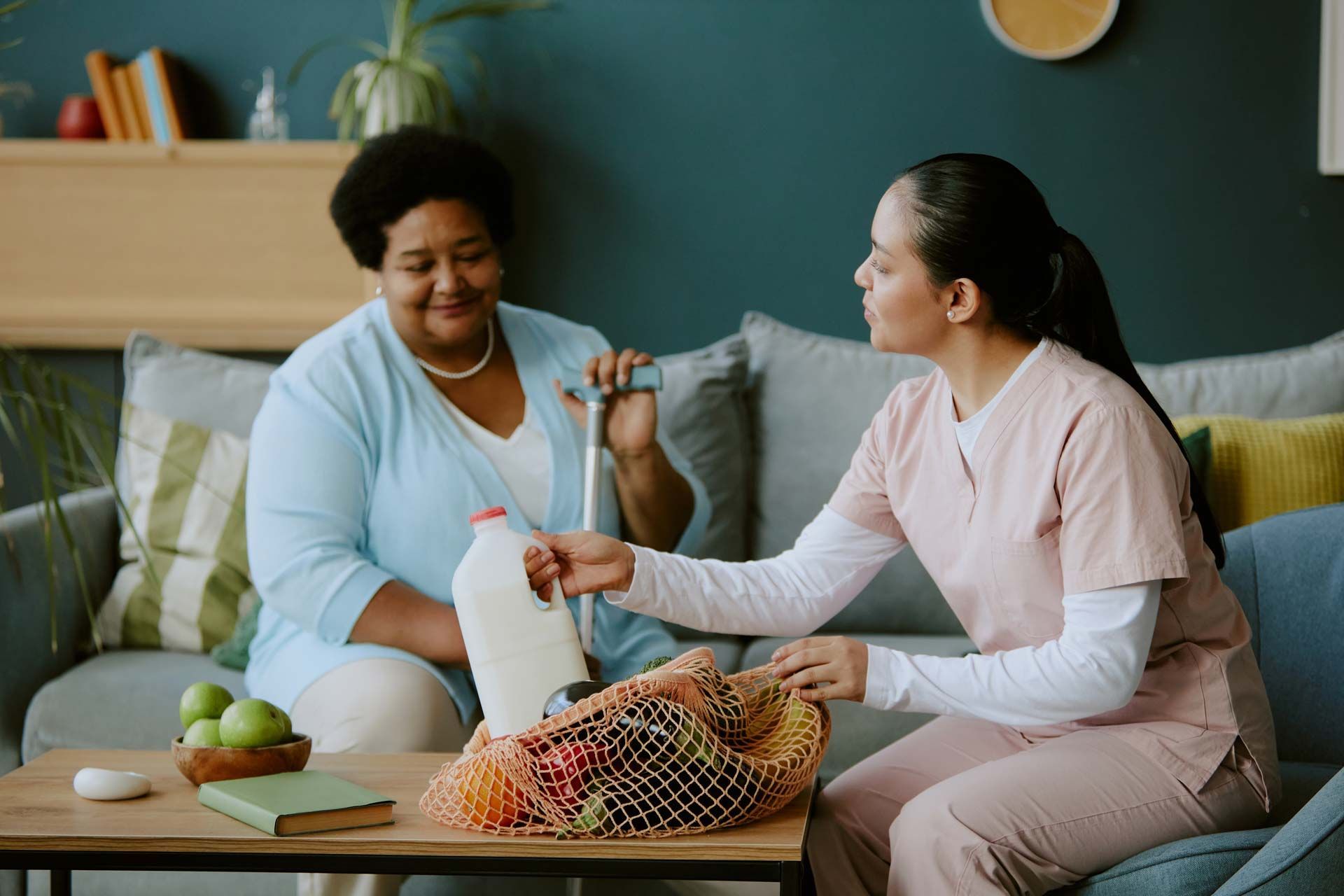 Woman assists a person seated on a couch with groceries, including a milk jug.