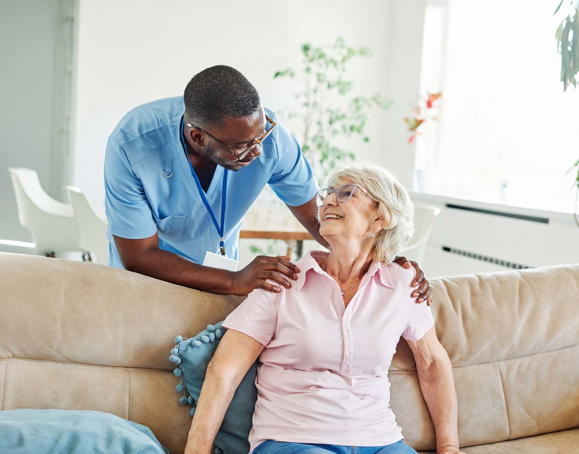 Woman in blue scrubs assisting an older woman in a pink shirt on a couch.