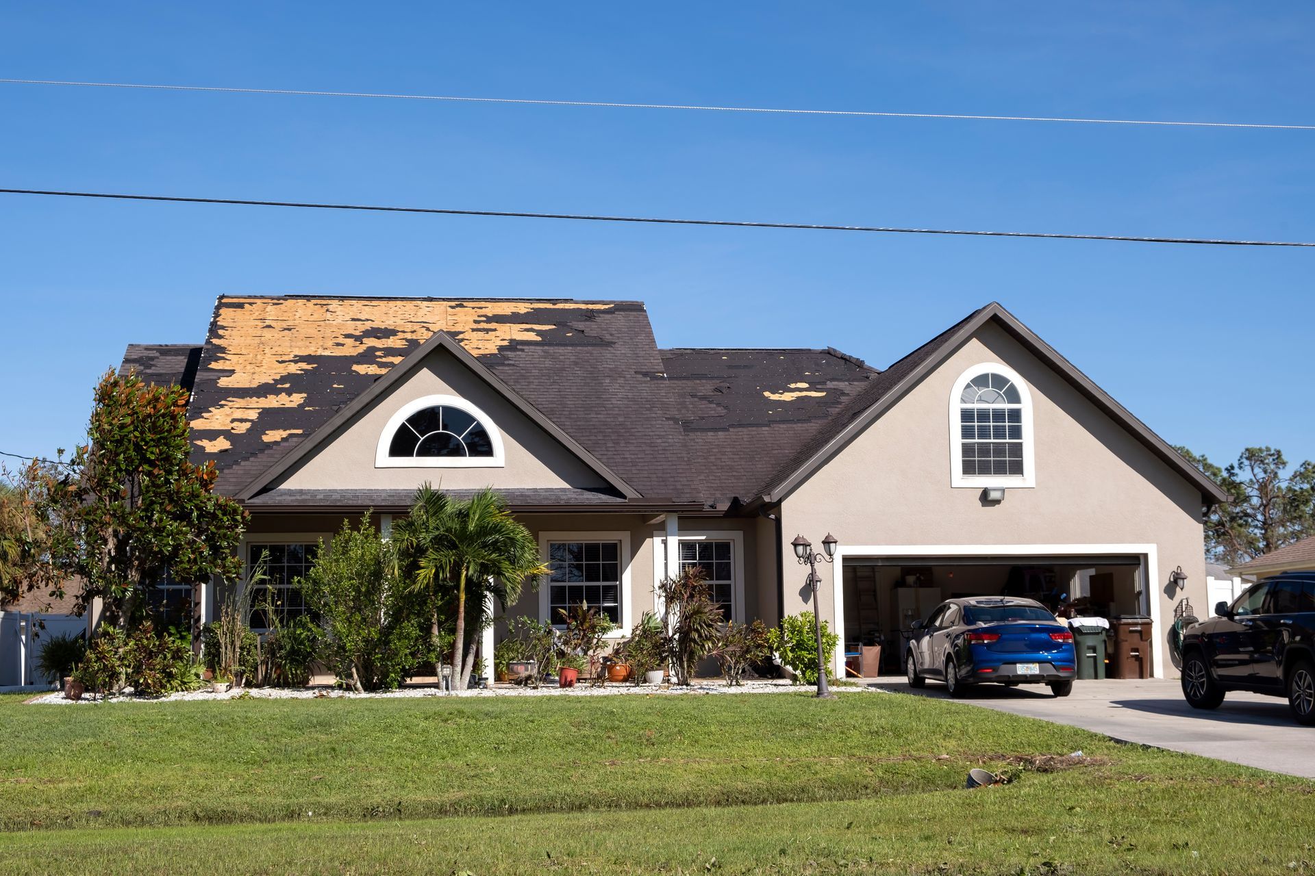 House with damaged roof and cars in the driveway. Green grass, blue sky.