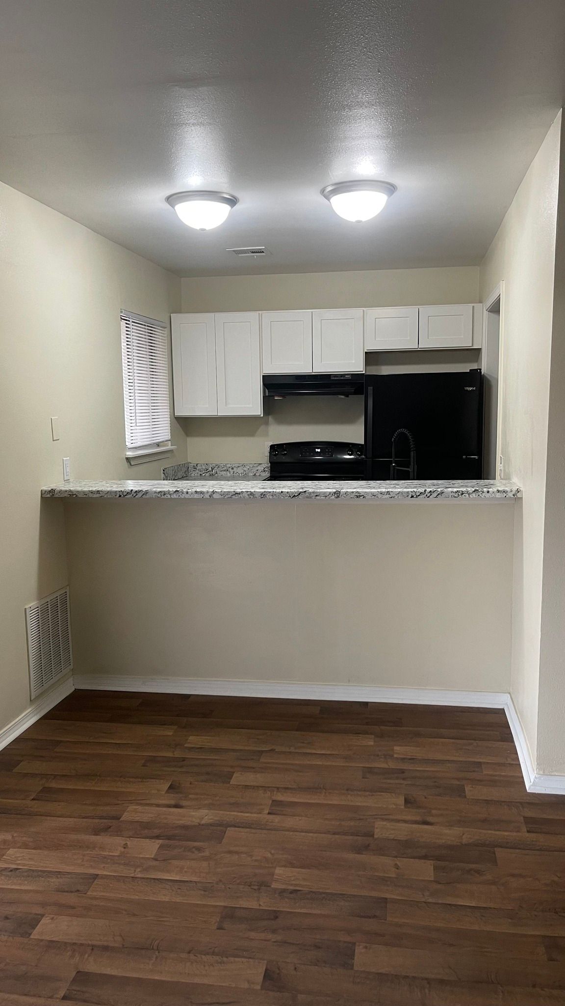 An empty kitchen with hardwood floors , white cabinets , a stove and a refrigerator.