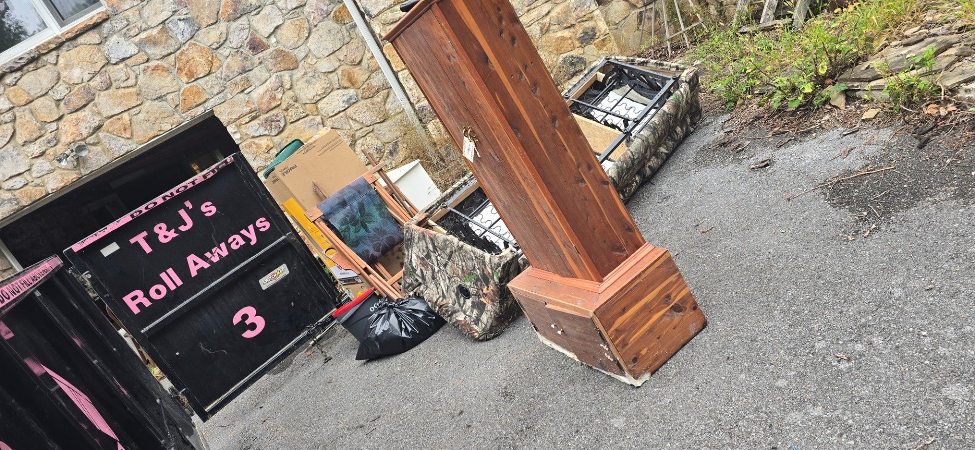 Pile of discarded items: a wooden post, trash bags, cardboard, and a dumpster. Outdoors on pavement.