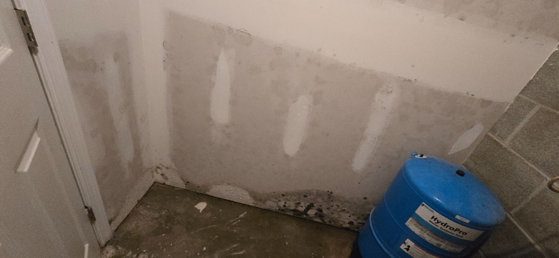 Basement corner with water tank, unfinished drywall, and concrete floor.