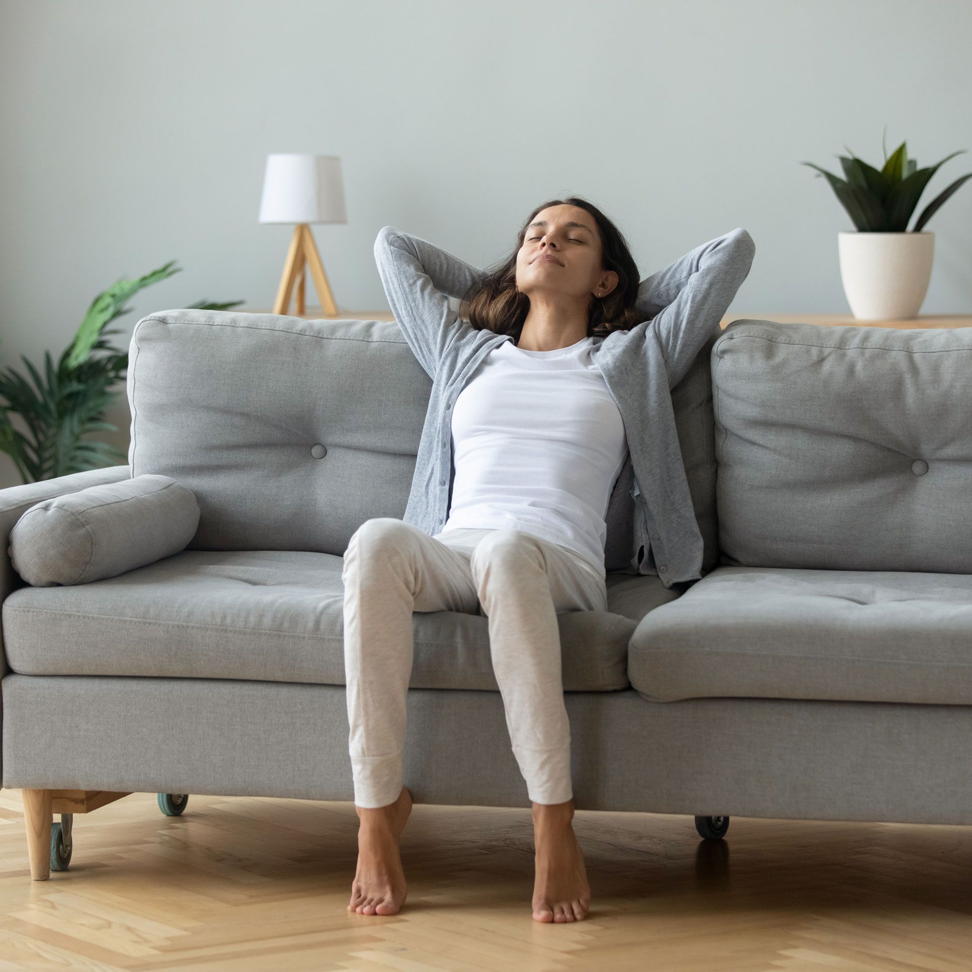 Woman relaxing on a gray sofa with hands behind her head, eyes closed.