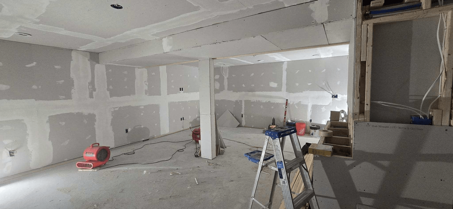Interior under construction: drywall installed on walls and ceiling, ladder, red blower fan.