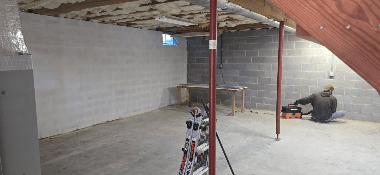 Basement construction: Person working near tools and support posts. Gray block walls, concrete floor.