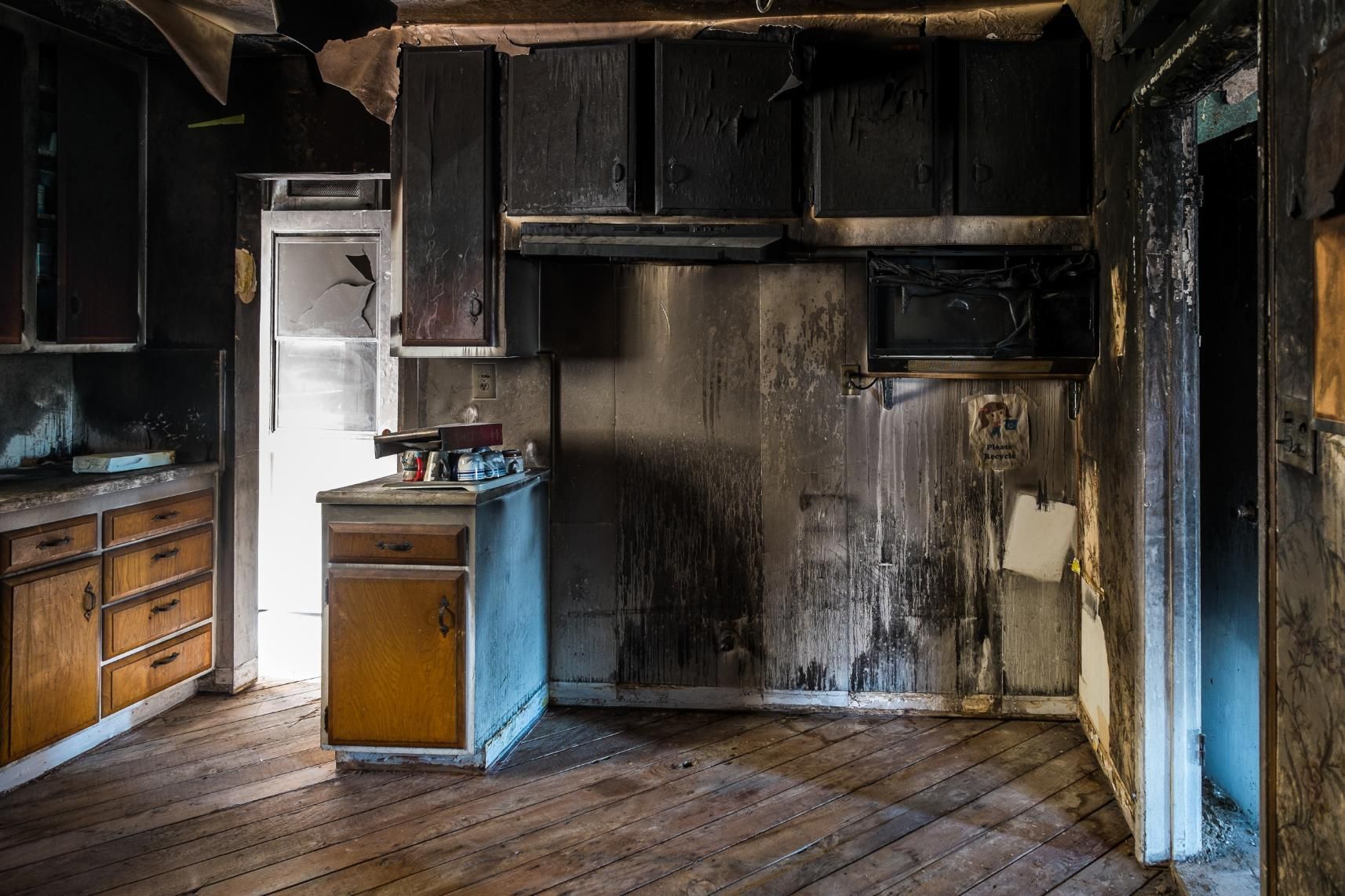 Charred kitchen interior after a fire; damaged cabinets, blackened walls, open door to light.