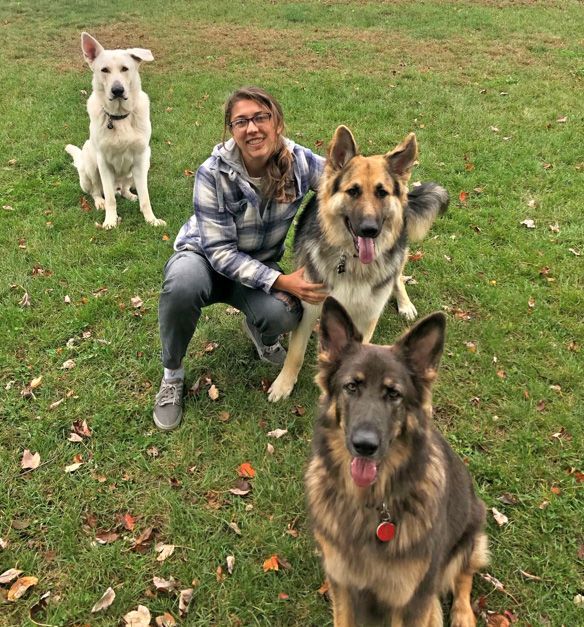 Laura is kneeling down next to two german shepherds in a grassy field.