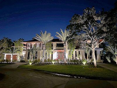 A large house at night with illuminated landscaping and trees, glowing with warm exterior lighting.