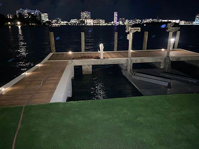 A night view of a lit wooden dock extending into dark water, with a blurred city skyline visible in the distance.