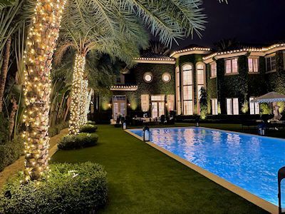 A luxury house with ivy-covered walls, illuminated by warm holiday lighting at night, next to a lit rectangular swimming pool.
