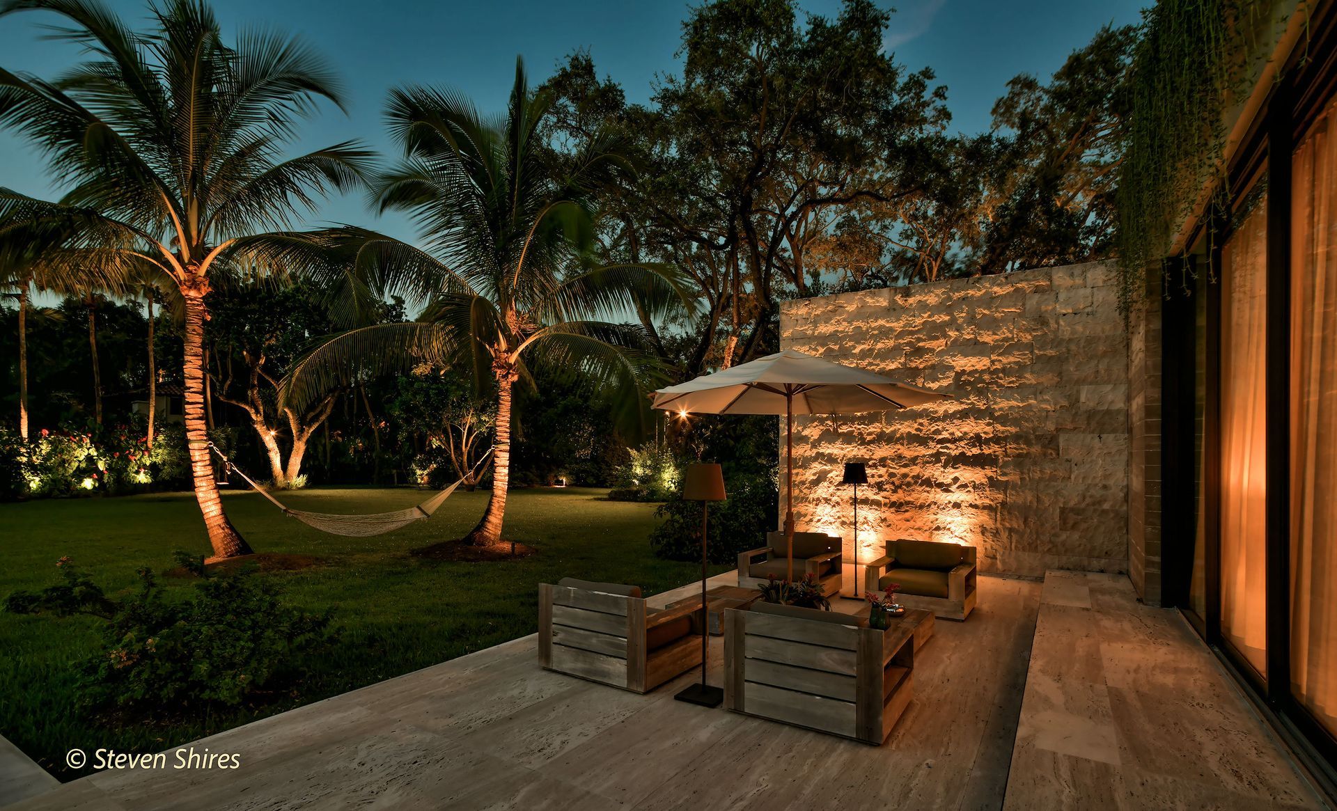 A patio with wooden chairs and an umbrella at dusk, featuring a stone wall, palm trees, and a grassy lawn.