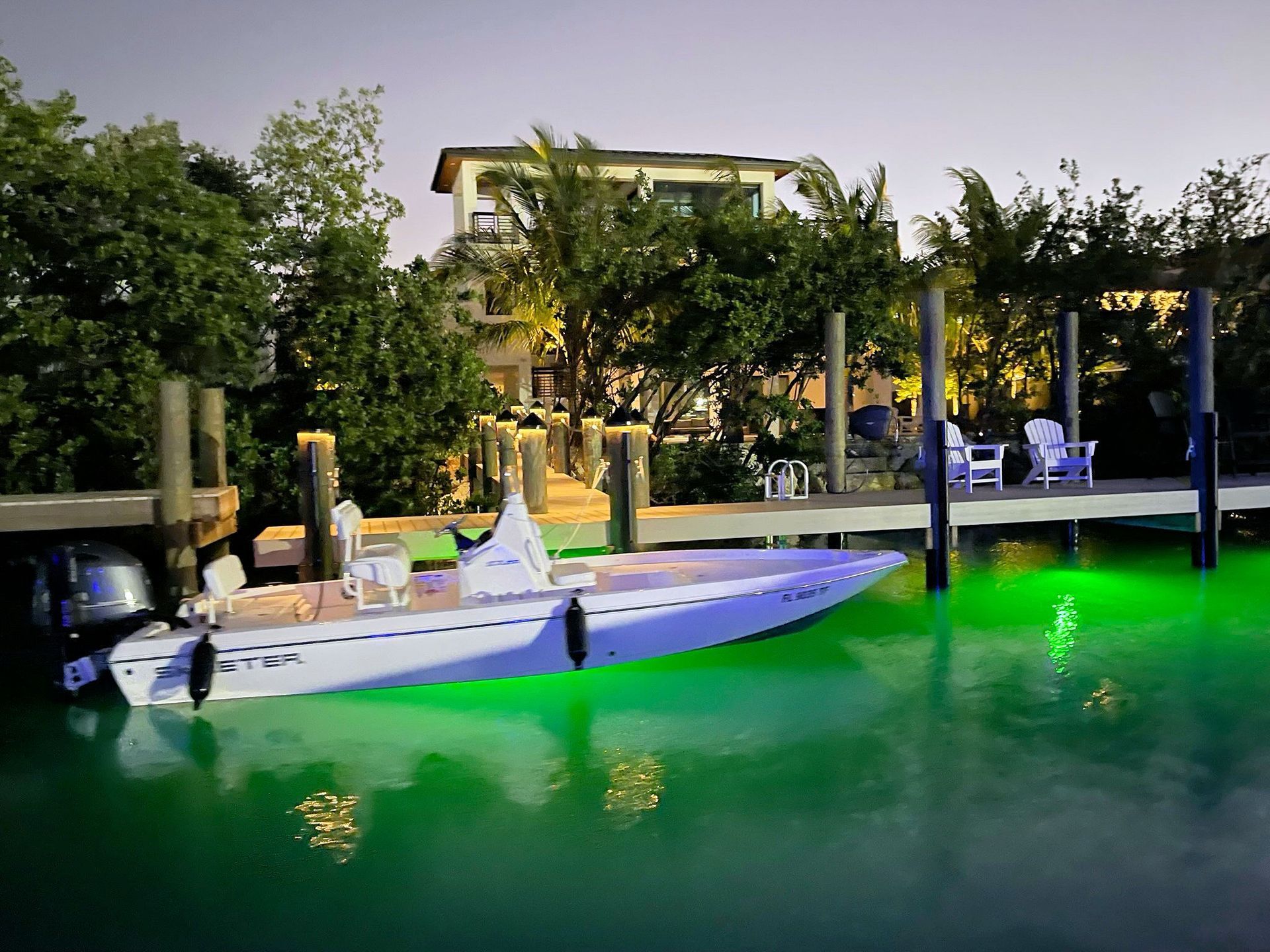 A white boat illuminated with glowing green underwater lights docked at a waterfront home at twilight.