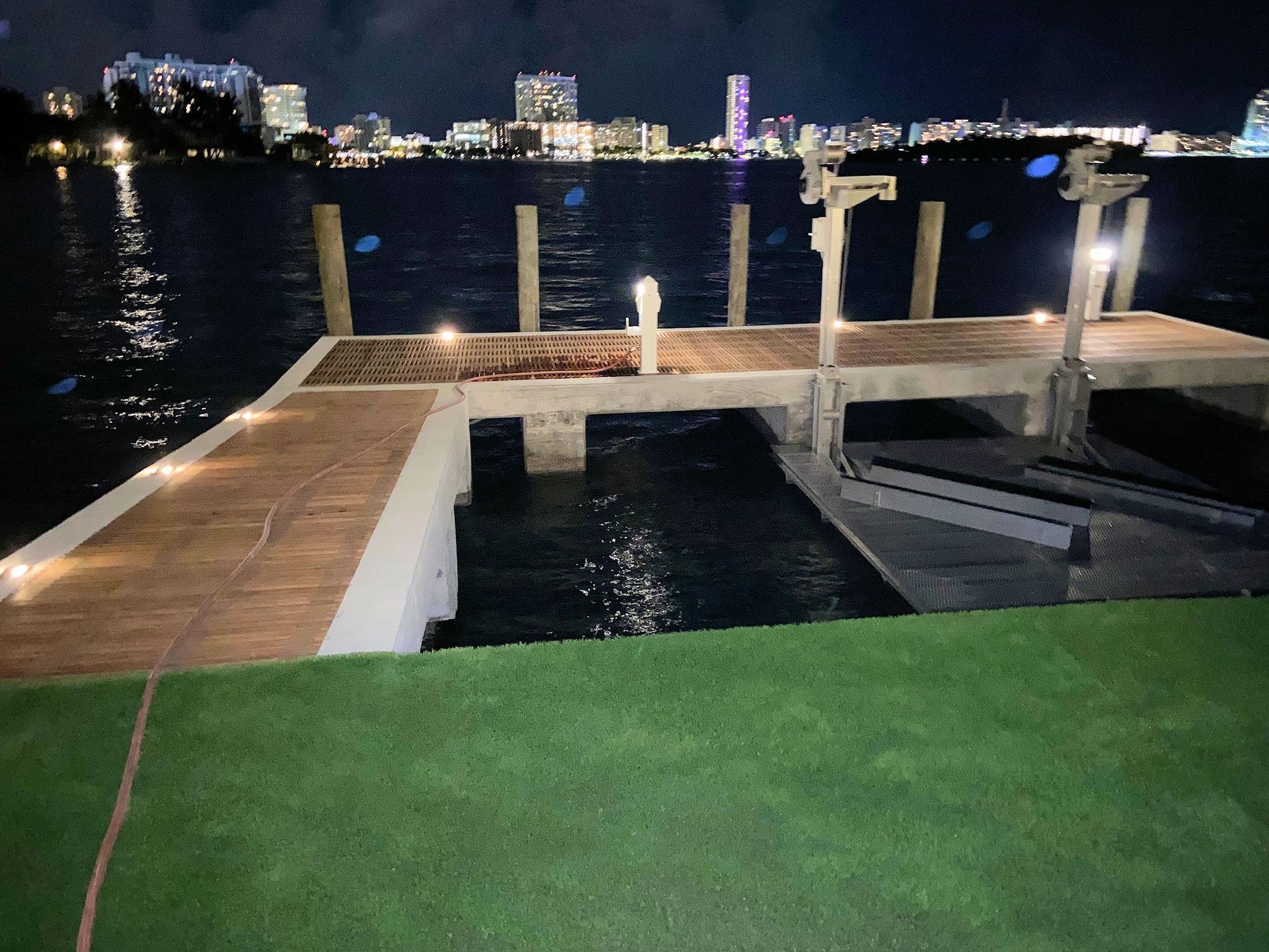 A cement dock with integrated lights extending into dark water at night, overlooking a distant, illuminated city skyline.