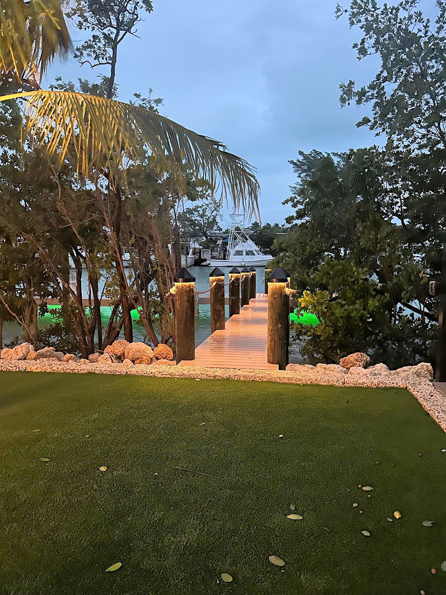 A wooden dock with illuminated posts extends from a lush green lawn toward a boat anchored in calm water at dusk.