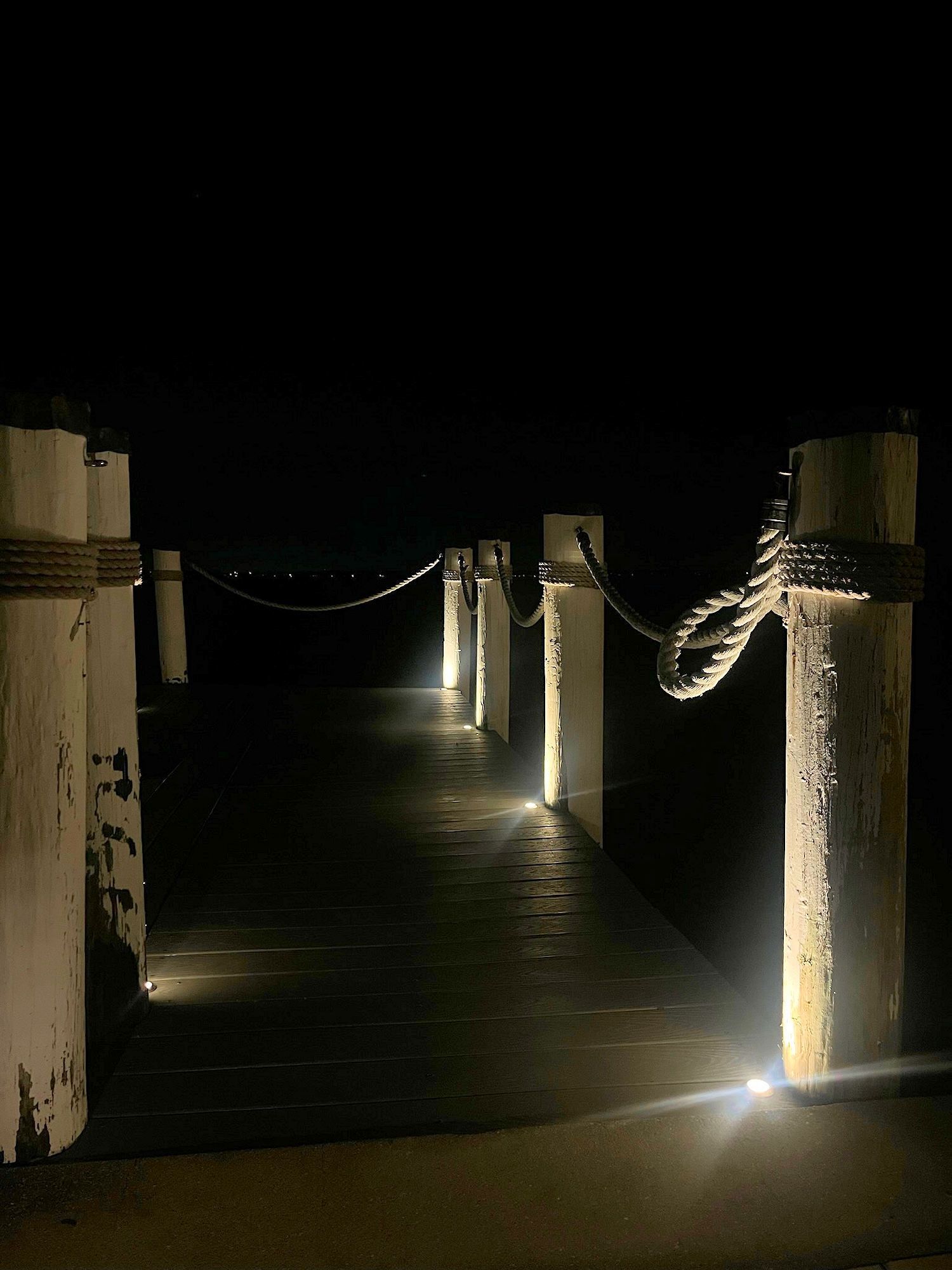 A wooden pier at night, illuminated by soft ground lights along white bollards connected by thick nautical ropes.