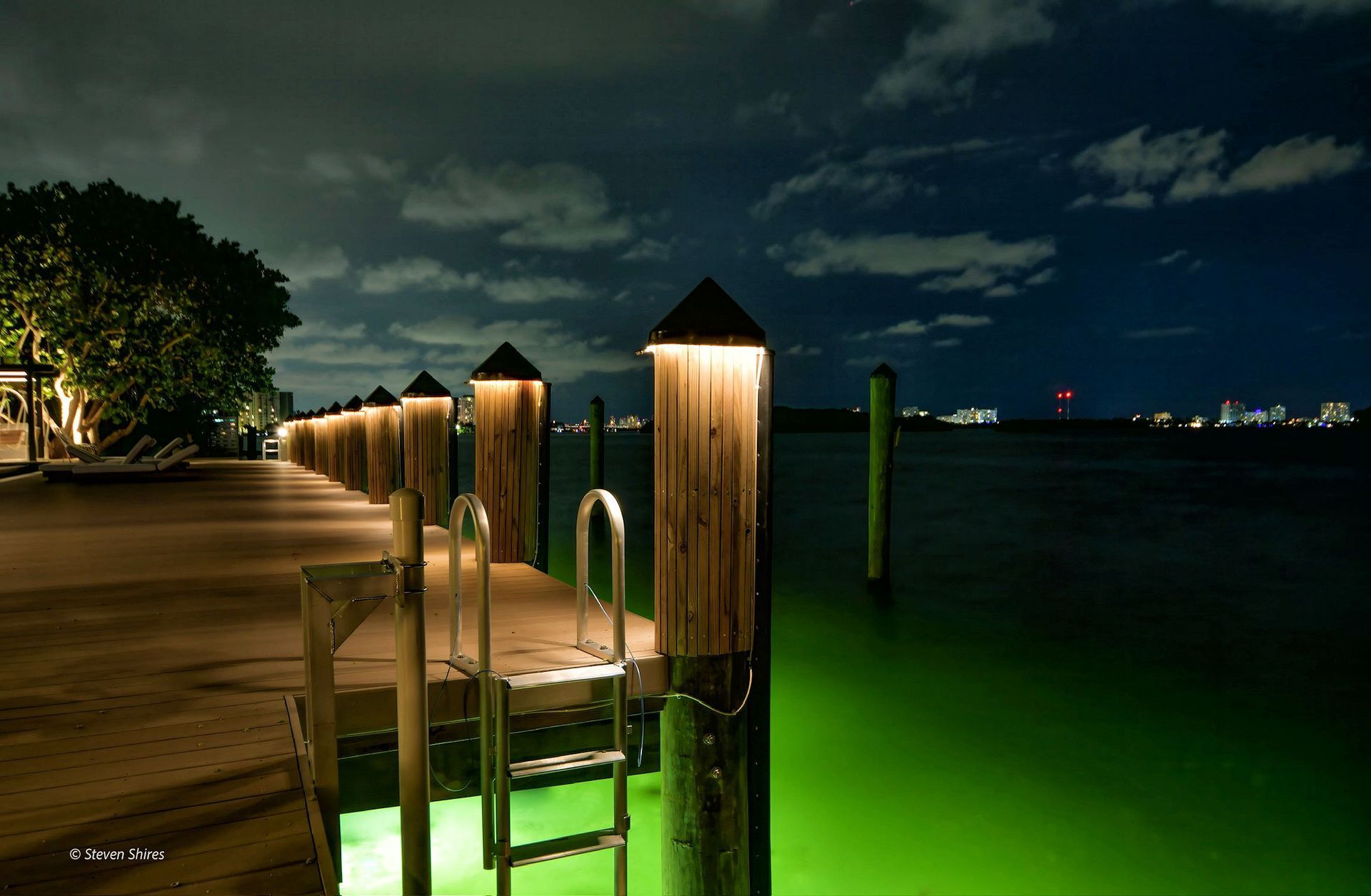 A wooden pier at night, lit by illuminated posts and glowing green water below, with a ladder leading into the sea.