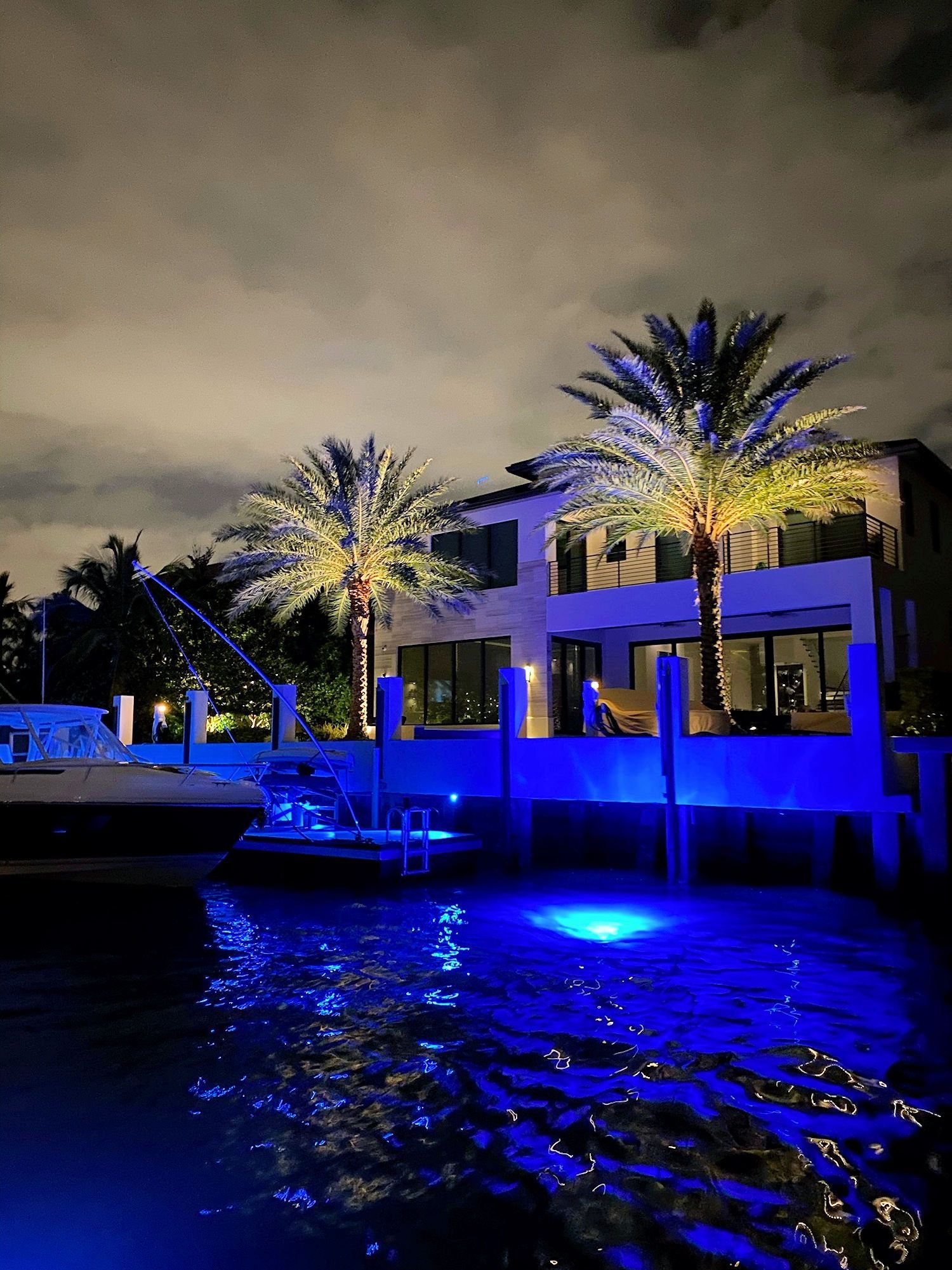 A two-story house at night with bright blue dock lighting reflecting on the water and two illuminated palm trees.