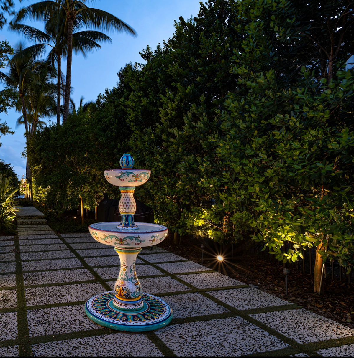 Lit garden walkway with a decorative two-tier fountain at dusk, surrounded by tropical greenery.