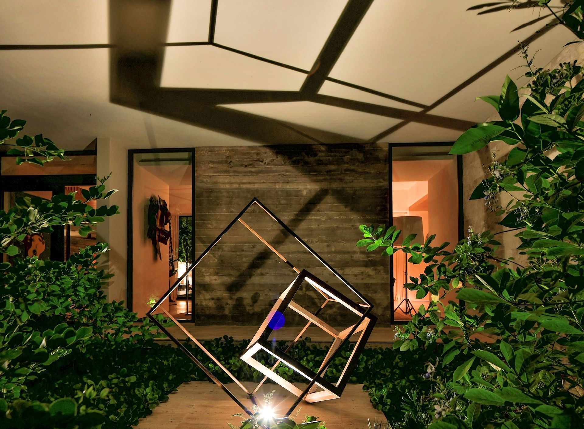 A geometric metal sculpture sits in a garden at night, casting angular shadows on the ceiling of a modern building.