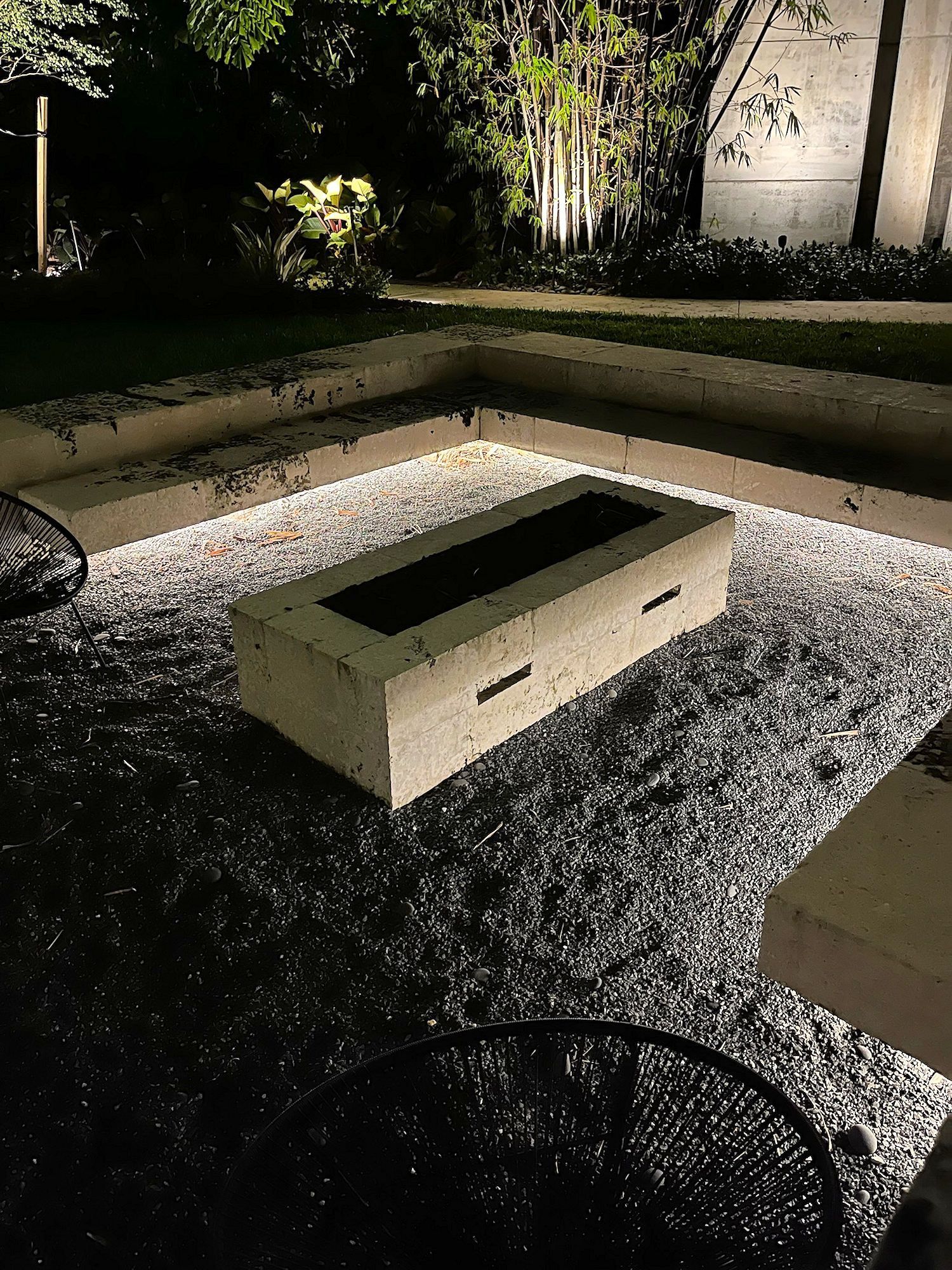 A rectangular concrete fire pit sits in a gravel-filled outdoor lounge area at night, illuminated by soft accent lighting.