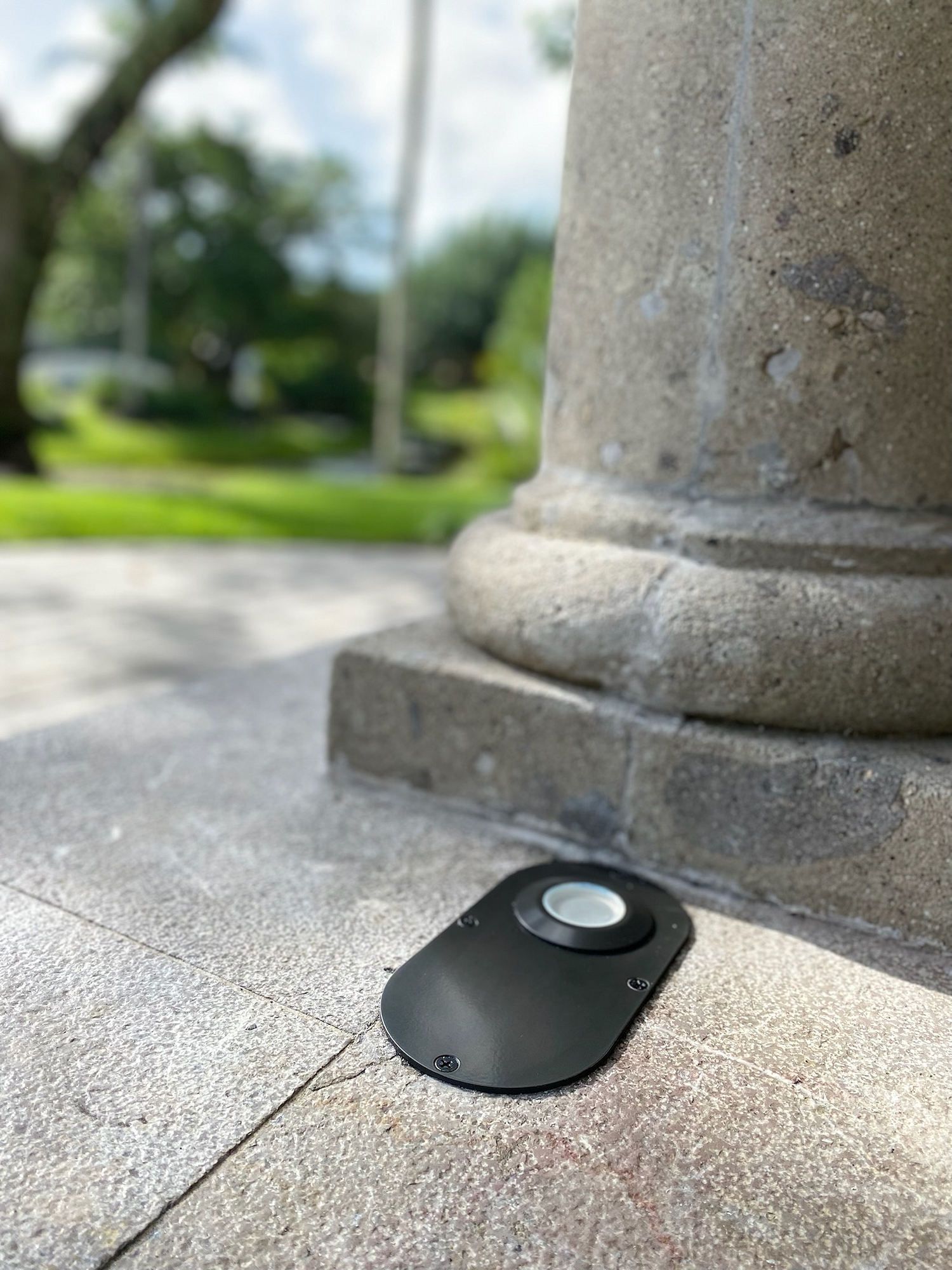A black, oval-shaped recessed landscape light fixture installed in a concrete patio next to a stone pillar.