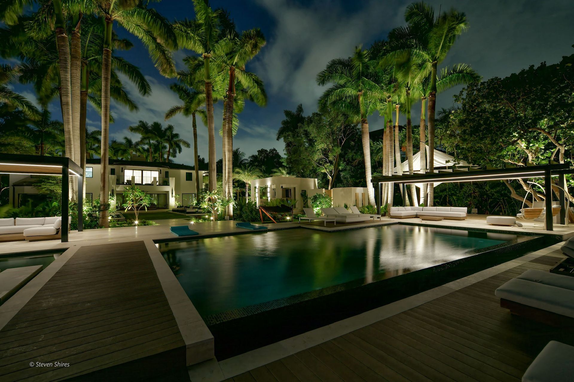 An illuminated luxury pool area at night, surrounded by lush palm trees and modern cabanas with outdoor seating.