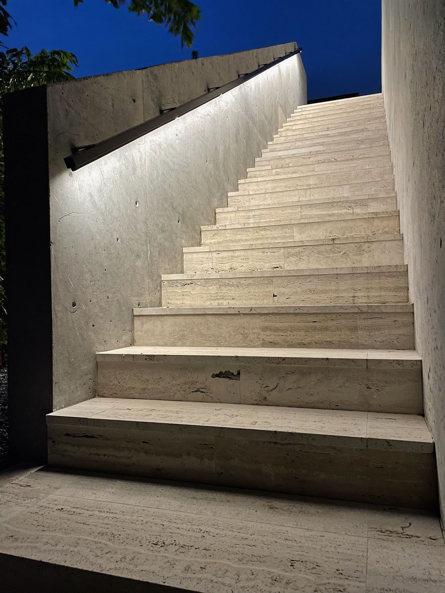 Modern stone staircase at night, illuminated by a handrail light against a textured concrete wall under a blue sky.