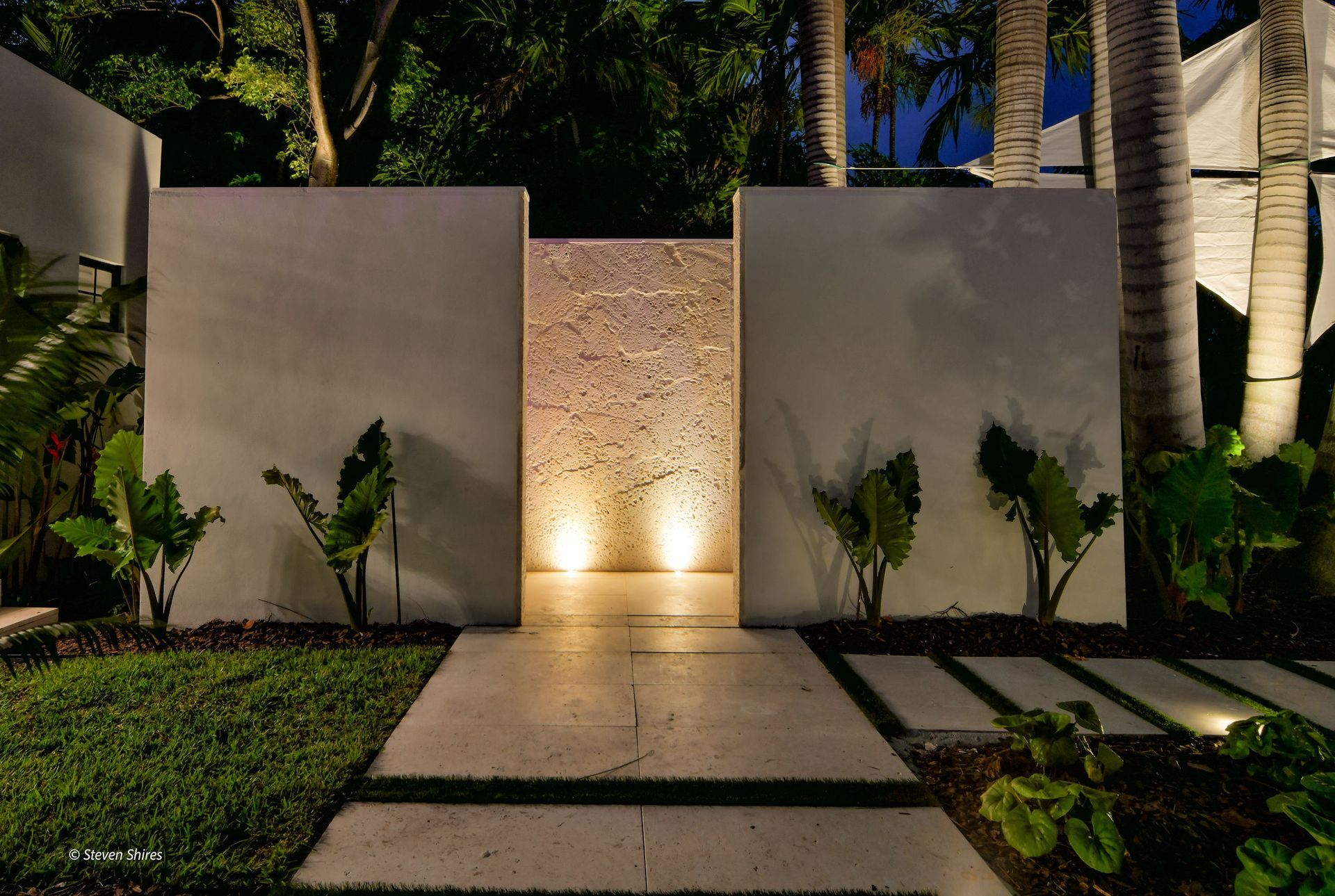 A lit outdoor stone pathway leads to a recessed, textured wall illuminated by warm uplighting between two white pillars.