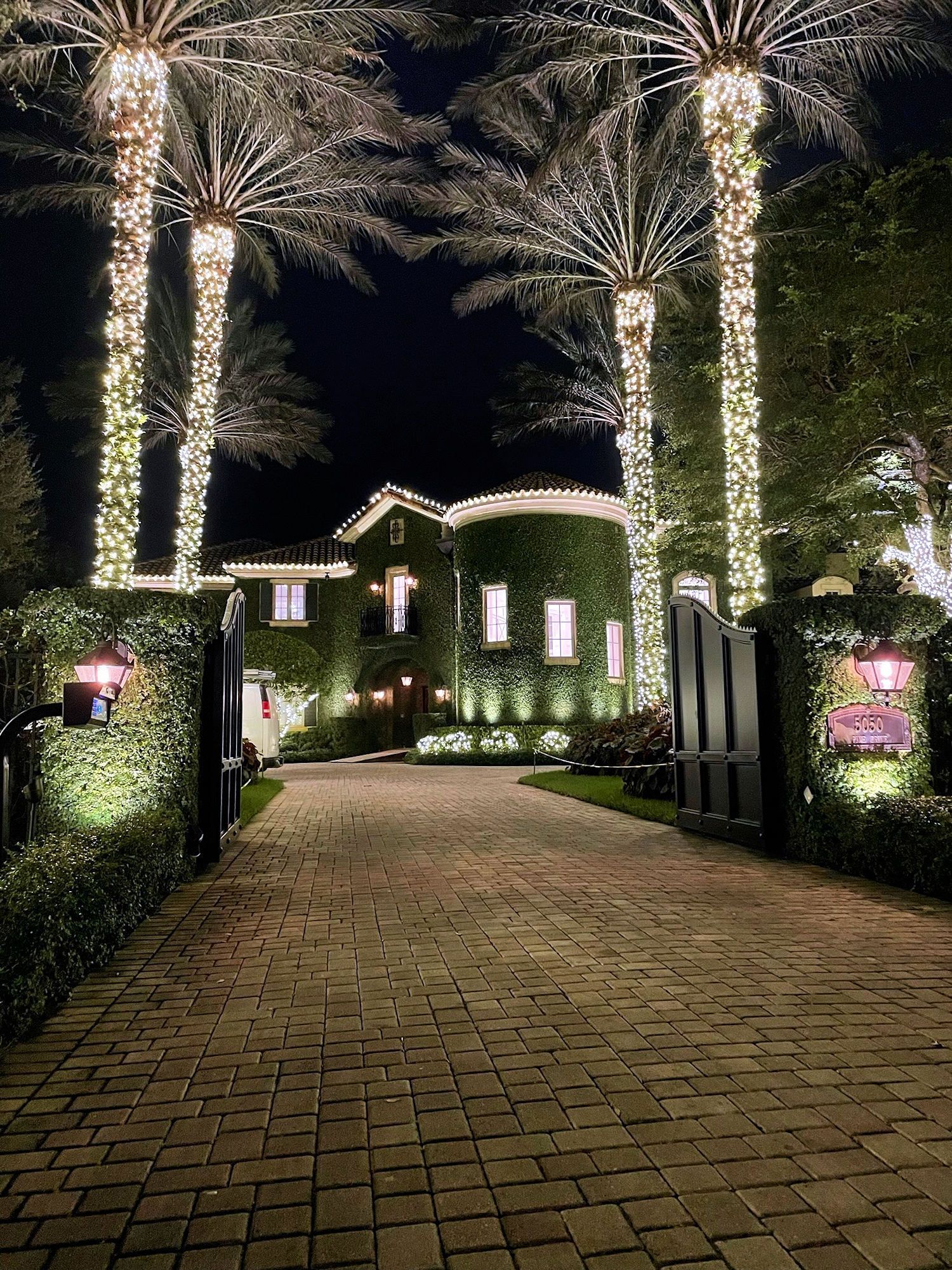 A cobblestone driveway leads to a large, ivy-covered home at night, framed by tall palm trees wrapped in white lights.