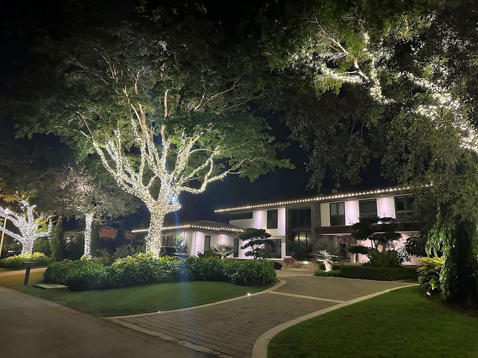 A luxurious two-story house at night with trees wrapped in white string lights and warm landscape lighting.